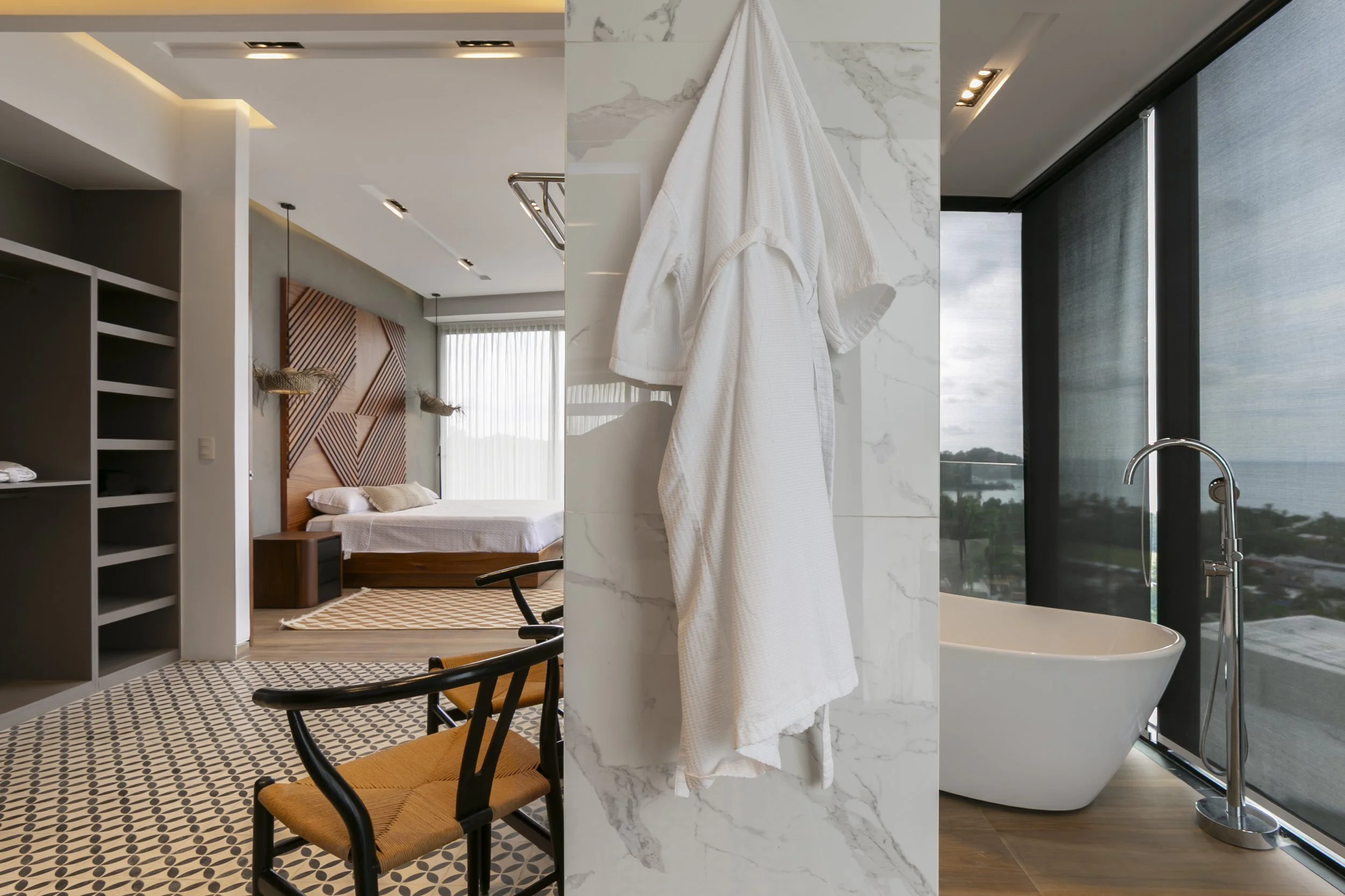 Interior view of a modern hotel room, featuring a bed with a wooden headboard, geometric wall art, seating area with chairs, and a marble wall with a hanging white bathrobe. A tub is visible next to floor-to-ceiling windows with a city view.