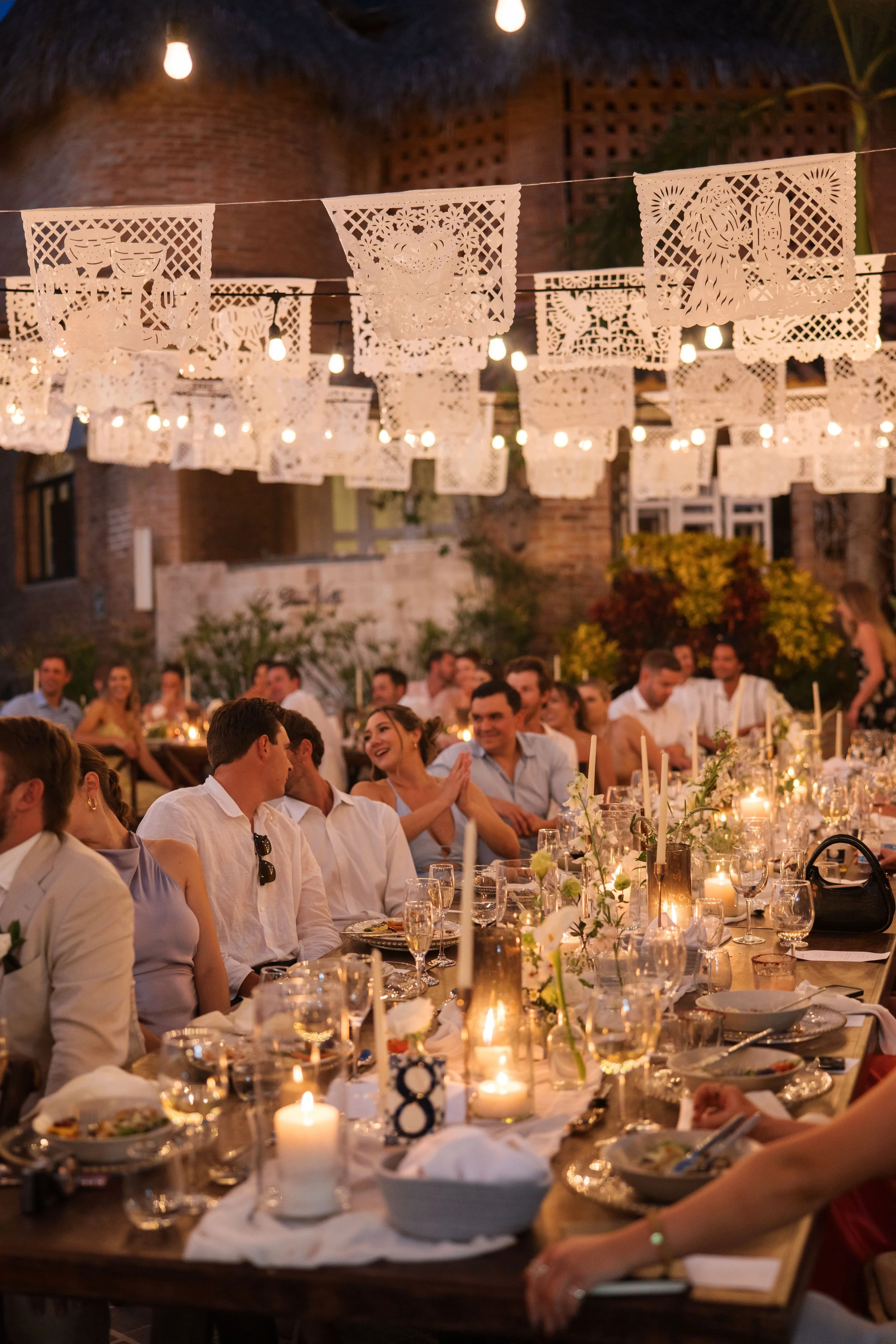 Outdoor evening dinner party with string lights, papel picado decorations, and a long table set with candles, glassware, and plates, attended by smiling guests.