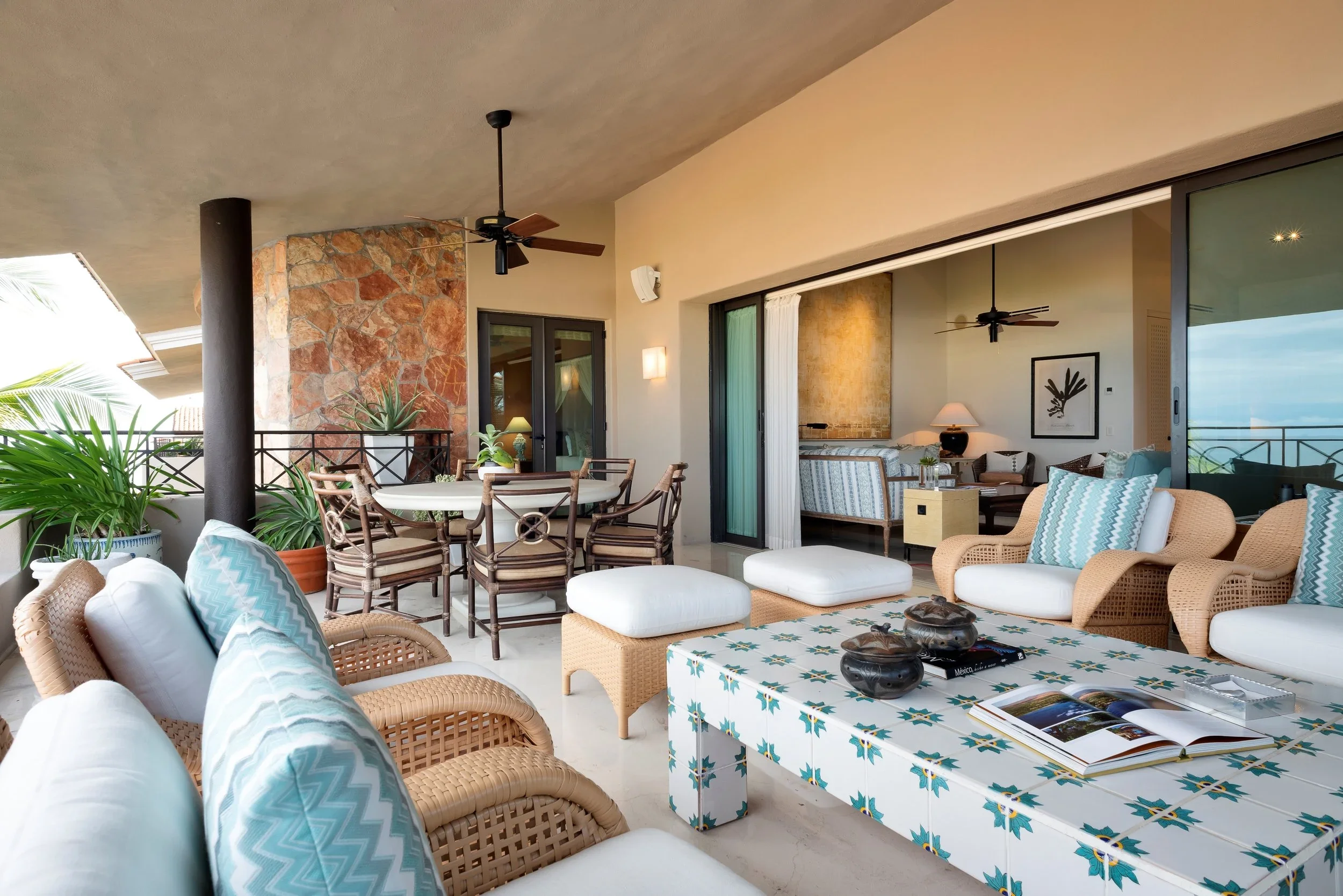 Open-air balcony with rattan chairs, a patterned table, potted plants, and a view of a neighboring building with a stone wall, overlooking the ocean.