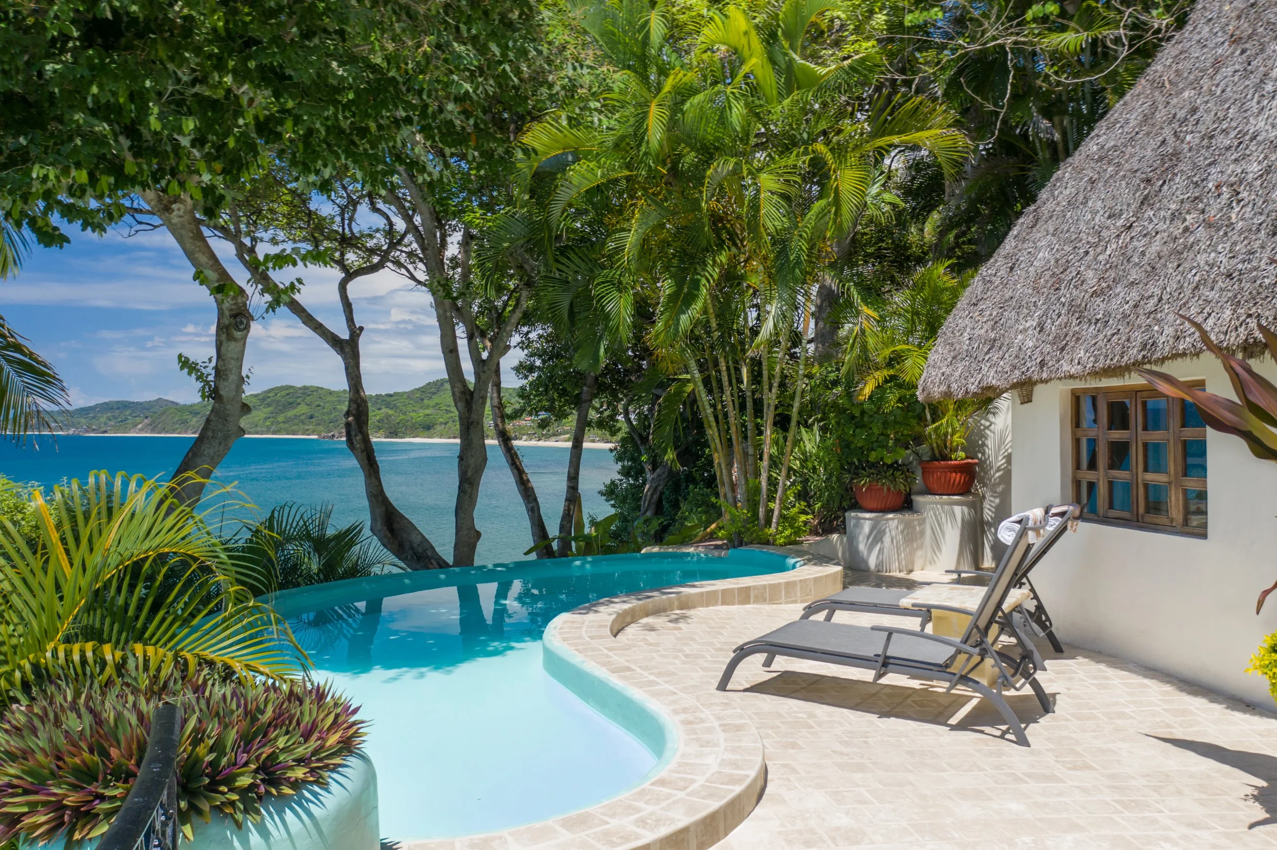 Luxury outdoor poolside area with lounge chairs beside a white house with a thatched roof, overlooking lush trees, a bay, and distant green hills.