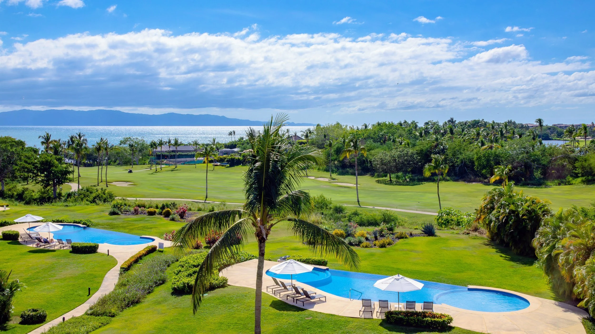 View of a tropical resort with a swimming pool, lounge chairs, umbrellas, lush green lawn, palm trees, a golf course, and the ocean in the background under a partly cloudy sky.