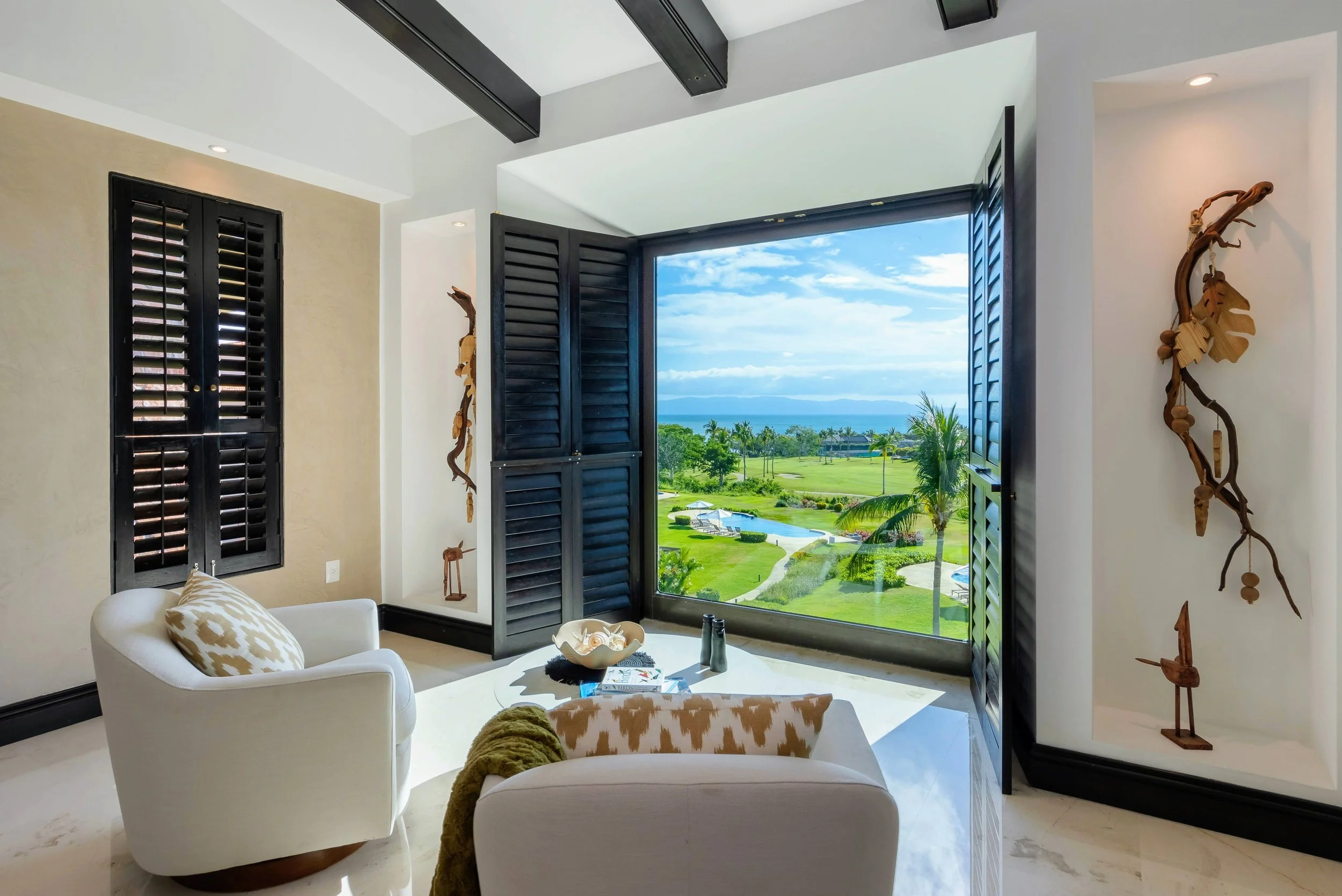 A living room with an open window showing a view of a golf course, palm trees, and the ocean, with black shutters and beige walls, decorated with tribal wooden art pieces.