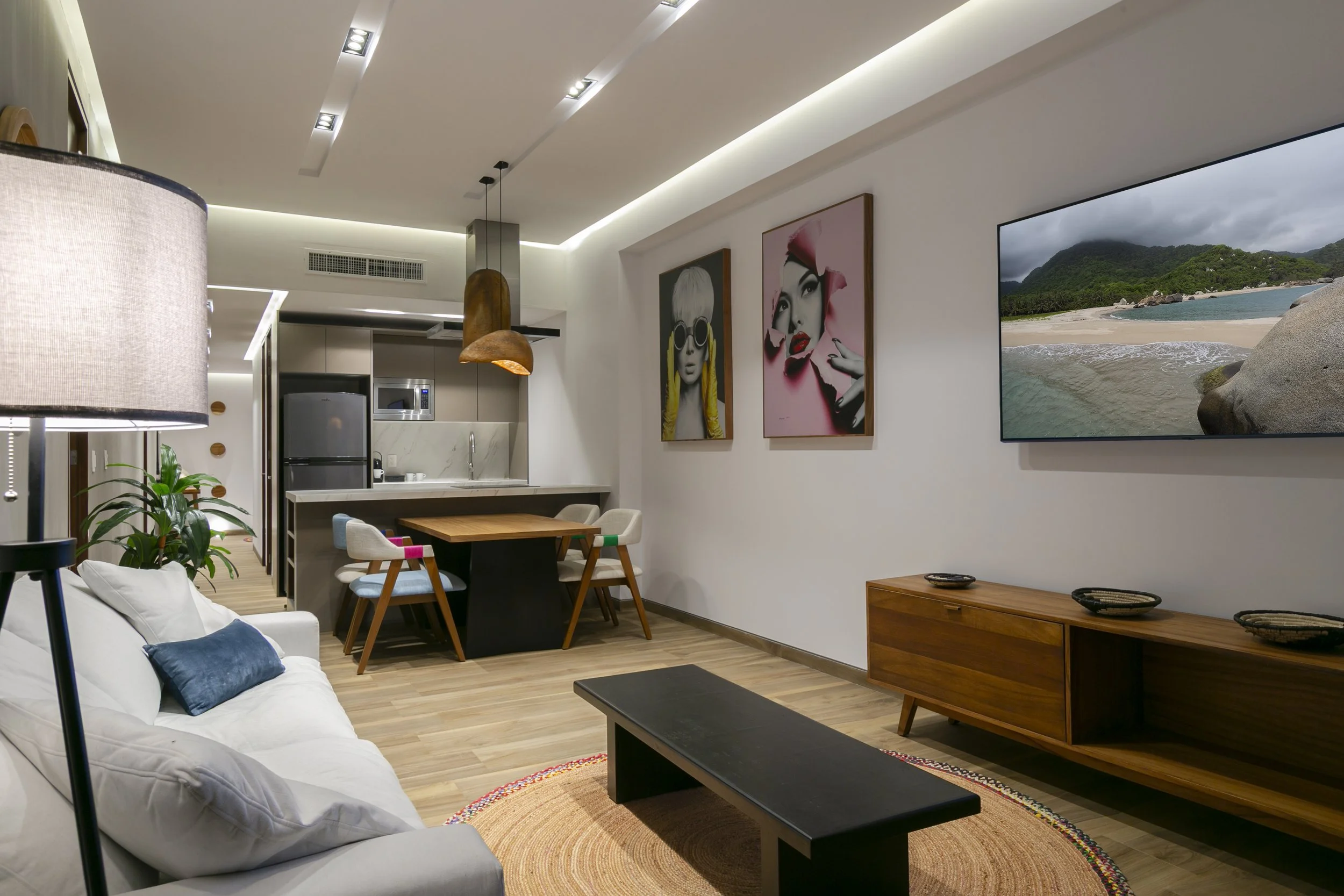 Modern living room with white sofa, coffee table, artwork on the wall, flat-screen TV, dining area, and a kitchen in the background.