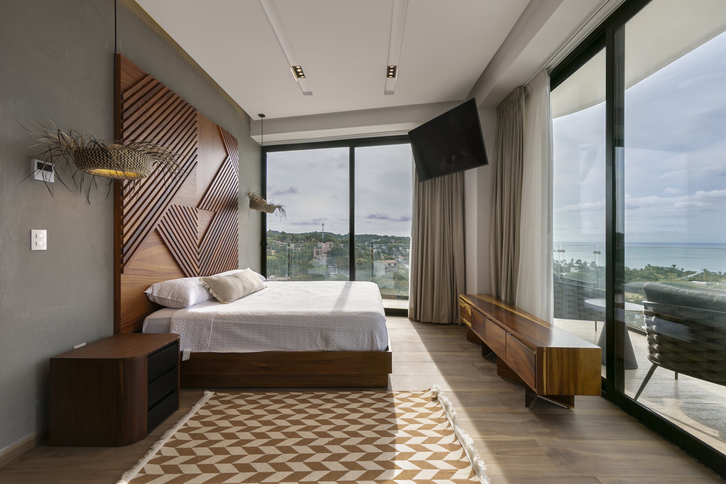 Modern bedroom with a large window view of the ocean, minimalist decor, wooden headboard with geometric design, wall-mounted lights, sliding glass door with curtains, wooden TV stand, and a patterned rug.