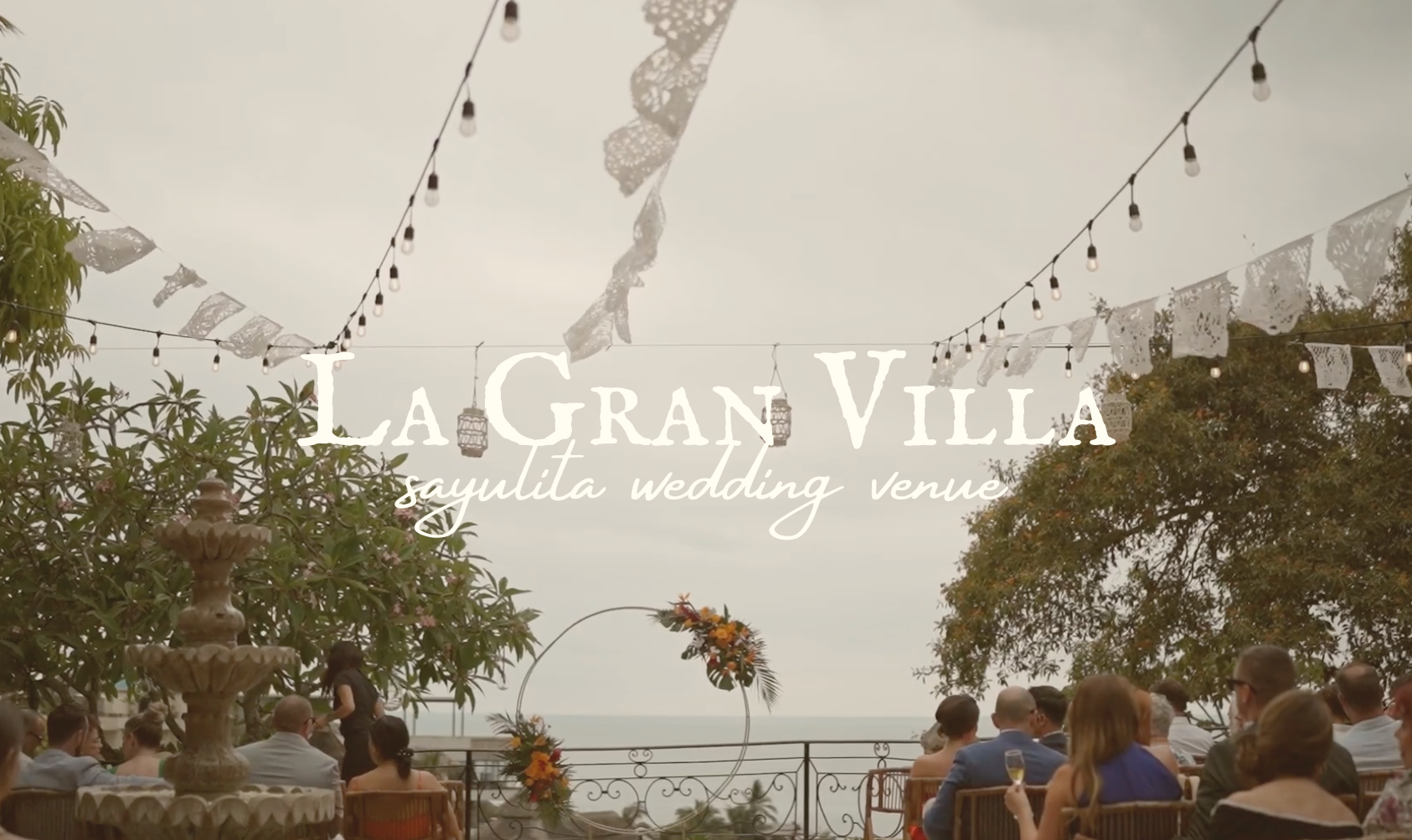 People seated outdoors at La Gran Villa Sayulita wedding venue during daytime, decorated with string lights, paper banners, lanterns, and floral arrangements near the ocean, with trees and cloudy sky in the background.