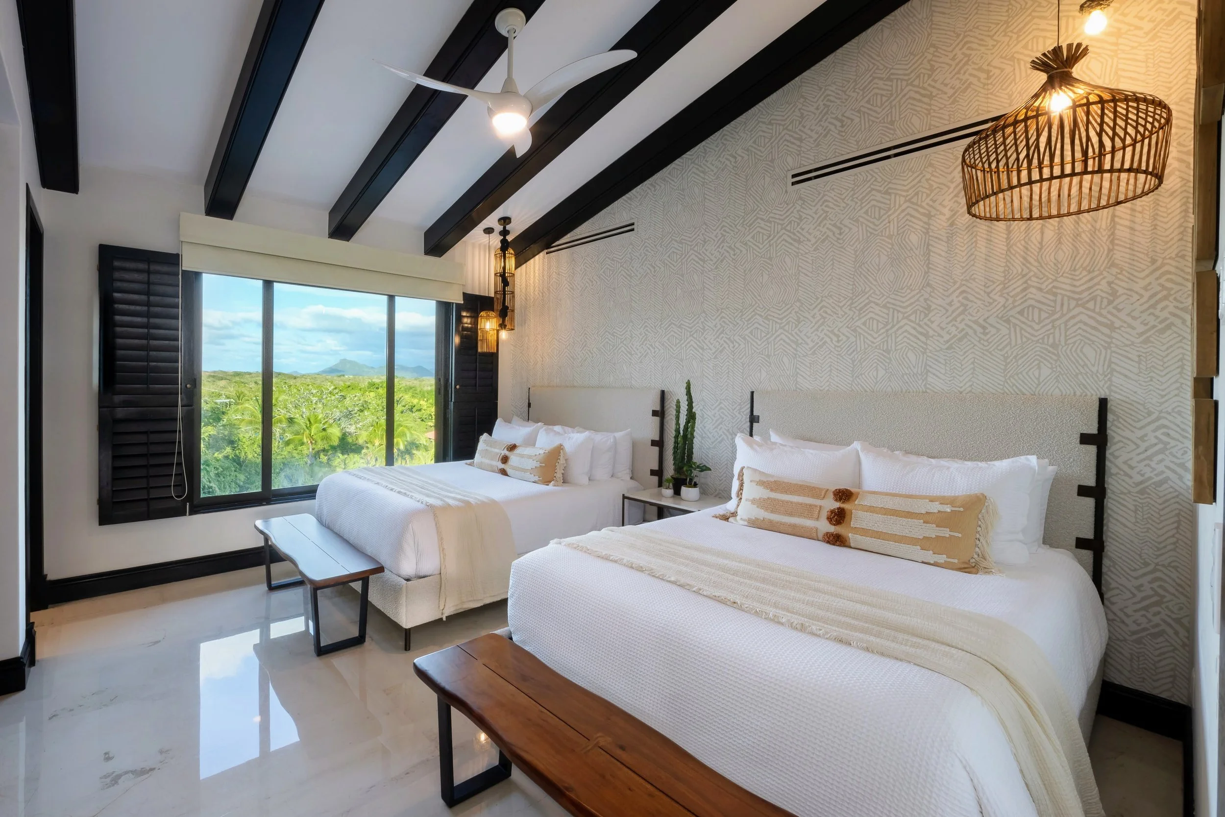 A bright hotel room with two large beds, white bedding, and decorative pillows. There is a window with black shutters showing a view of green trees and mountains. The room has black wooden beams on the ceiling, a ceiling fan, and hanging woven light fixtures. A small bench is at the foot of one bed, and a nightstand with a potted cactus and other decorations is between the beds.