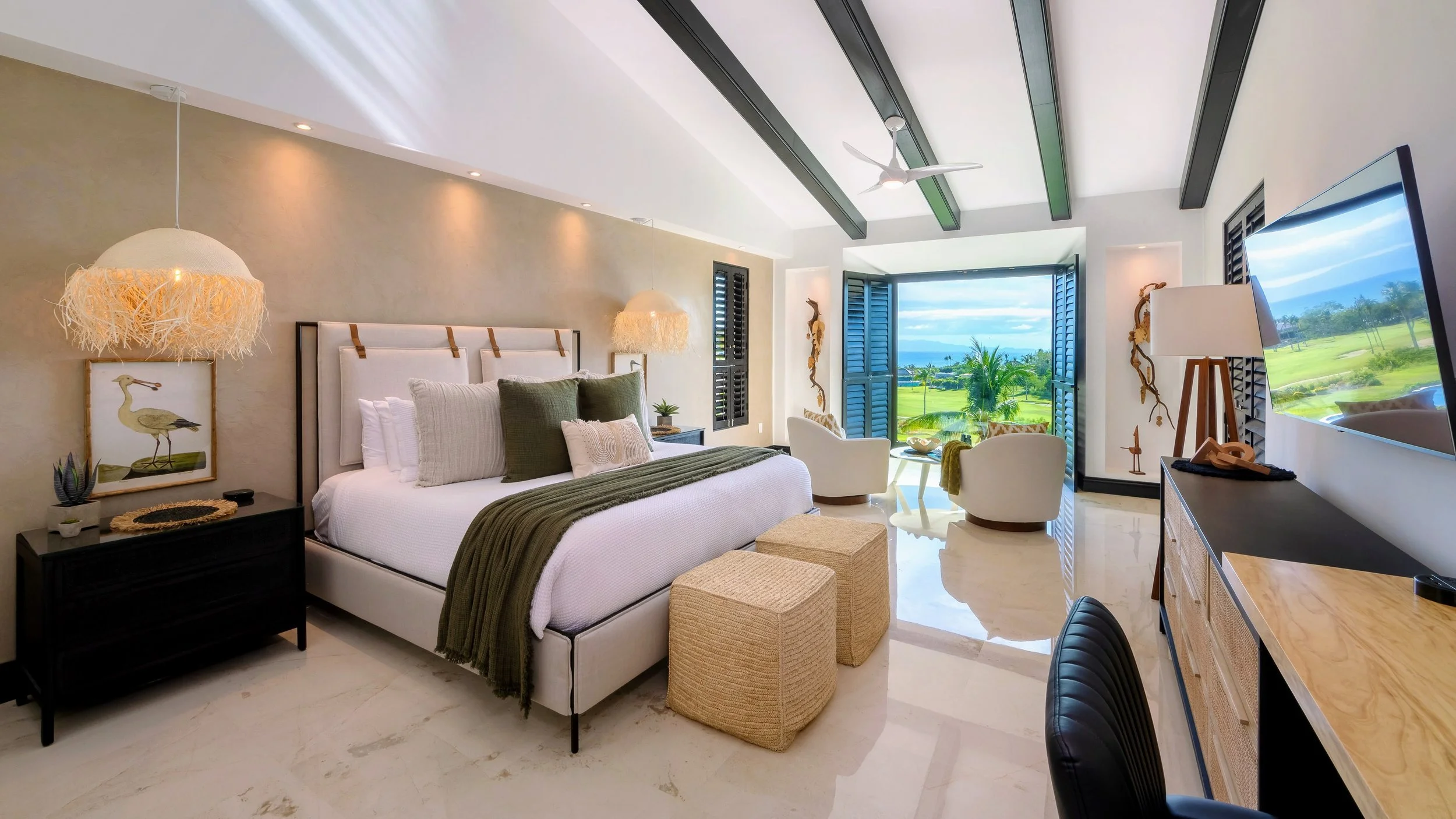 A bright, airy bedroom with a large bed decorated with white, beige, and green pillows and a green throw blanket. There are two woven ottomans in front of the bed, a black nightstand with plant and artwork, and a cream-colored wall with overhead spotlights. A desk with a lamp and a TV on the wall faces the bed. Large open windows with blue shutters reveal a view of a golf course and ocean in the distance.