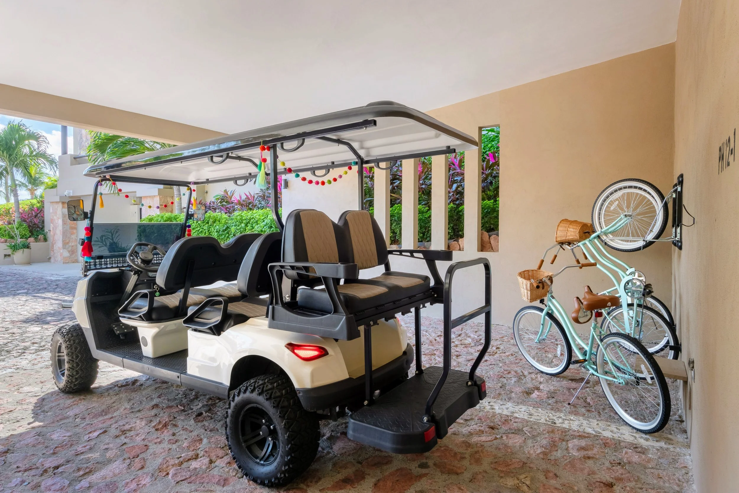 A golf cart parked in a covered outdoor area, decorated with colorful tassels and pom-poms, alongside two bicycles, one with a front basket, against a background of greenery and flowering plants.