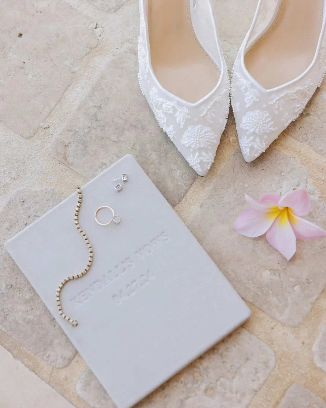 White bridal shoes with embroidery, a white jewelry box with pearl necklace, ring, and earrings, a pink flower, and a white guestbook with embossed text.