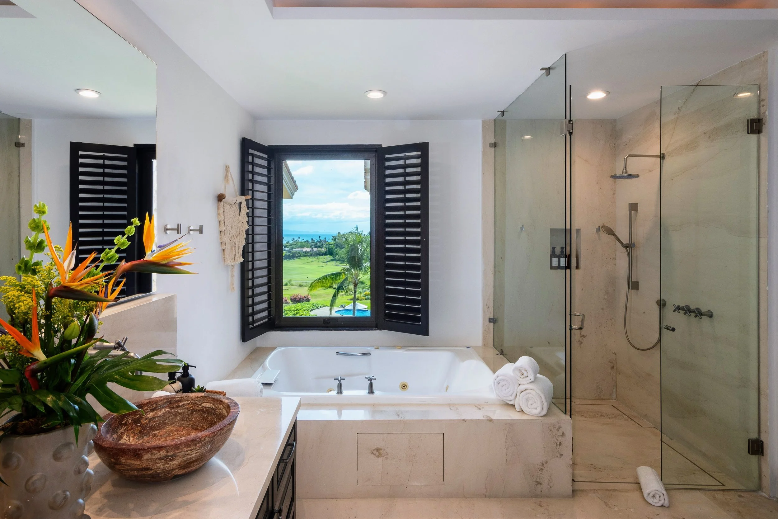 Luxury bathroom with a large bathtub, black window shutters opening to a scenic view of greenery and the ocean, a glass-enclosed shower, and a marble countertop with a wooden bowl sink and a flower arrangement.