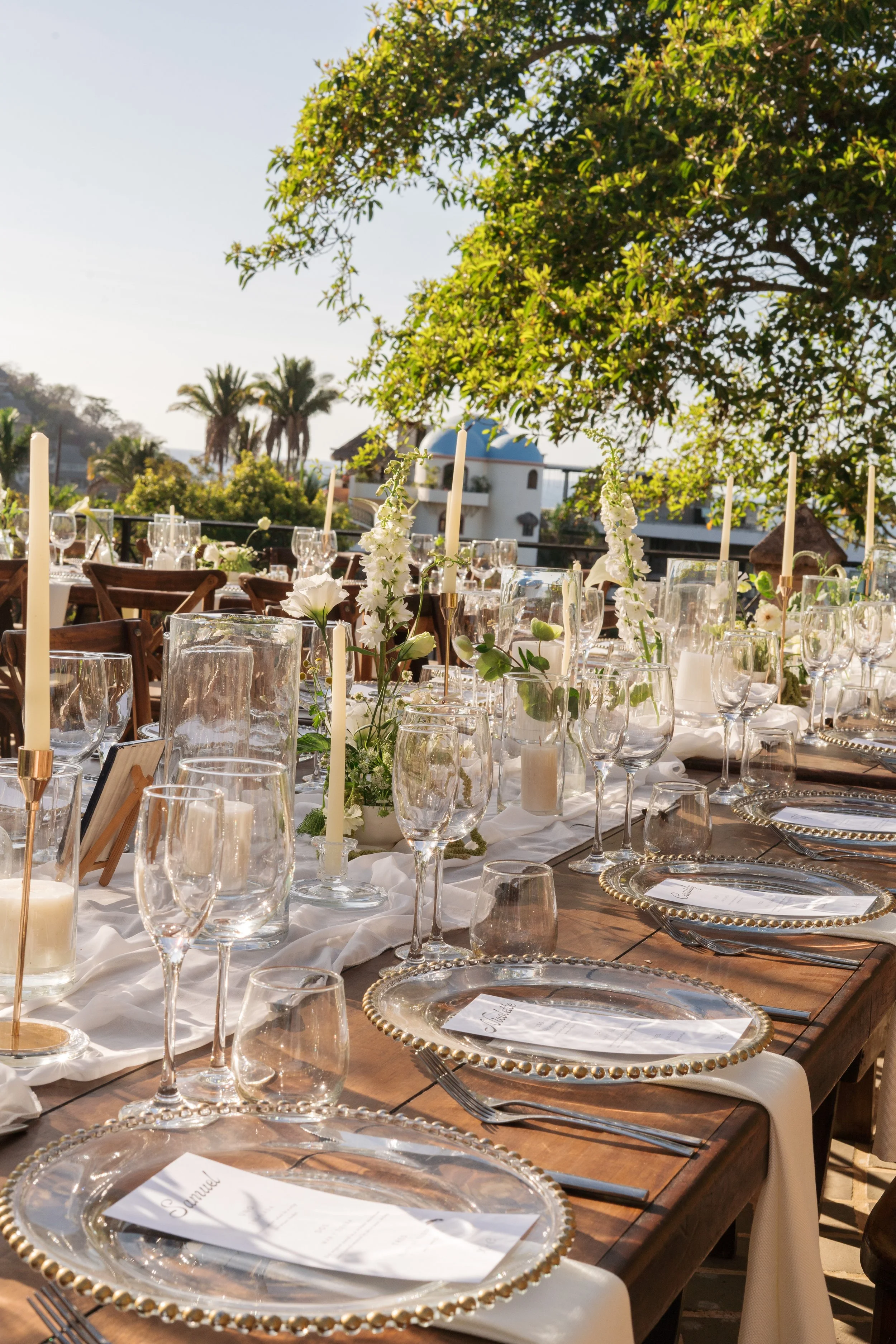 Elegant outdoor table setting with glass plates, wine glasses, candles, and white floral arrangements, under a tree with a blue-domed building in the background.