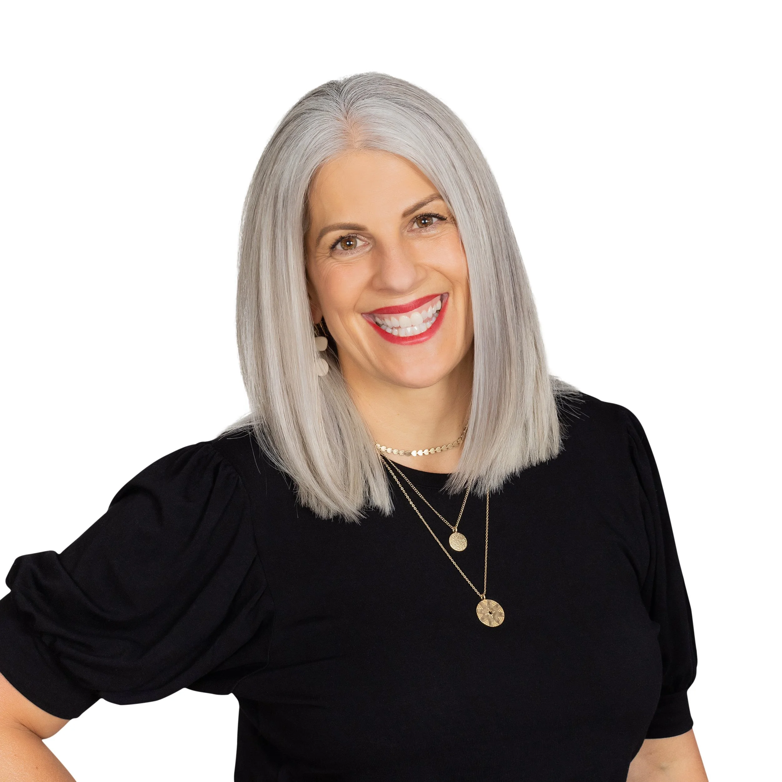 A smiling woman with shoulder-length gray hair, wearing a black top with puffed sleeves, layered necklaces, and earrings, posed against a plain white background.