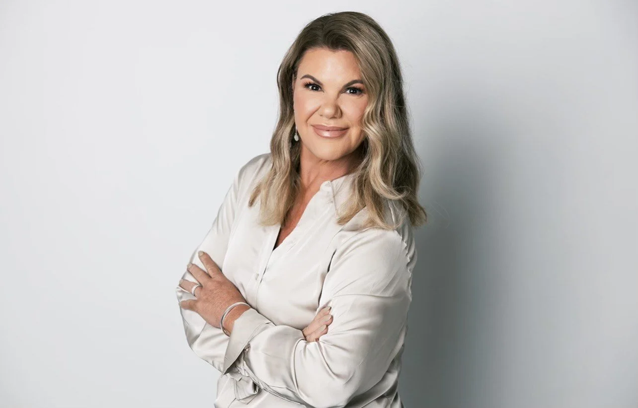 A woman with shoulder-length wavy blonde hair, wearing a white silk blouse, standing with arms crossed, smiling confidently in front of a plain light gray background.