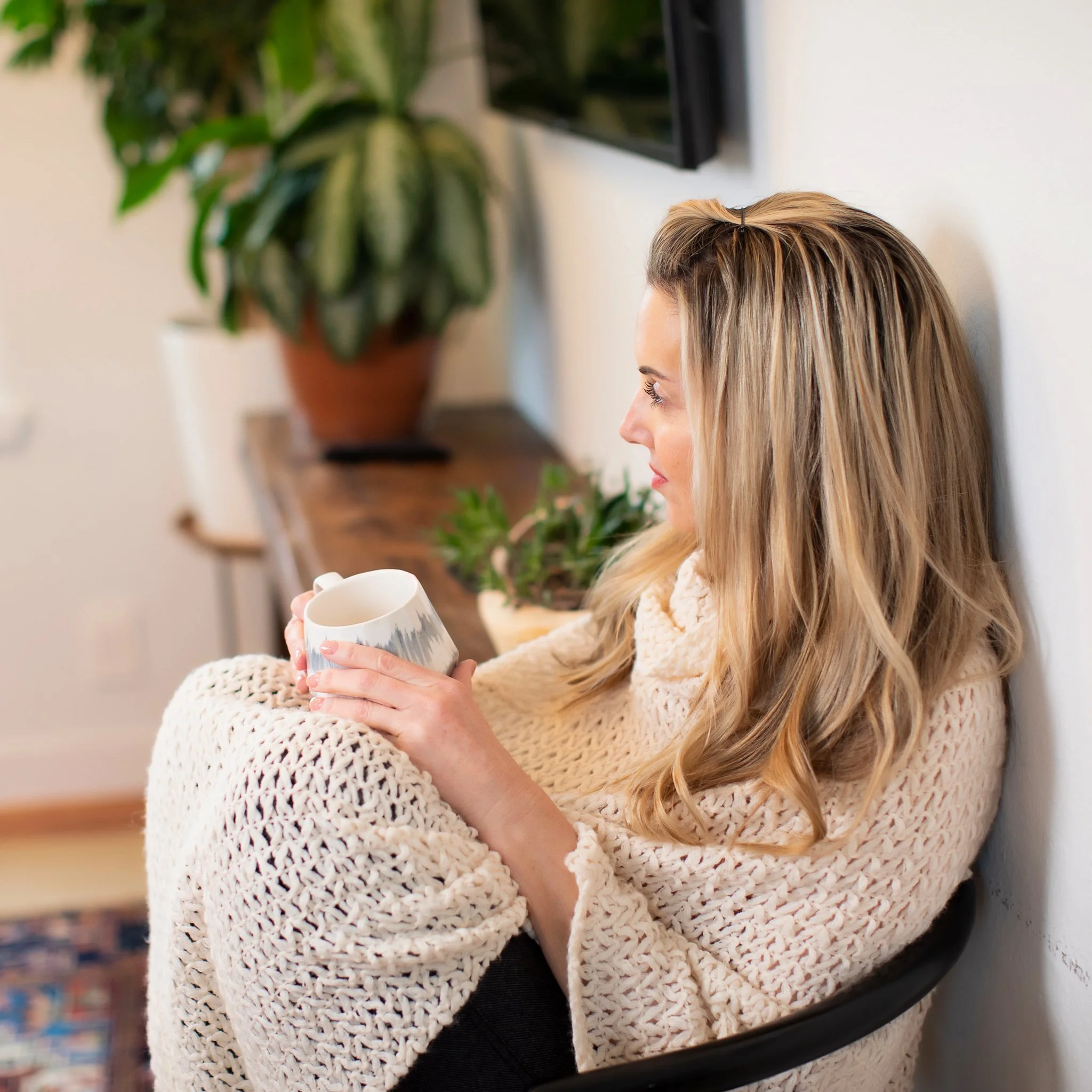 A woman with blonde wavy hair, wearing a cream knit sweater, sitting in a chair and holding a mug, looking thoughtful.