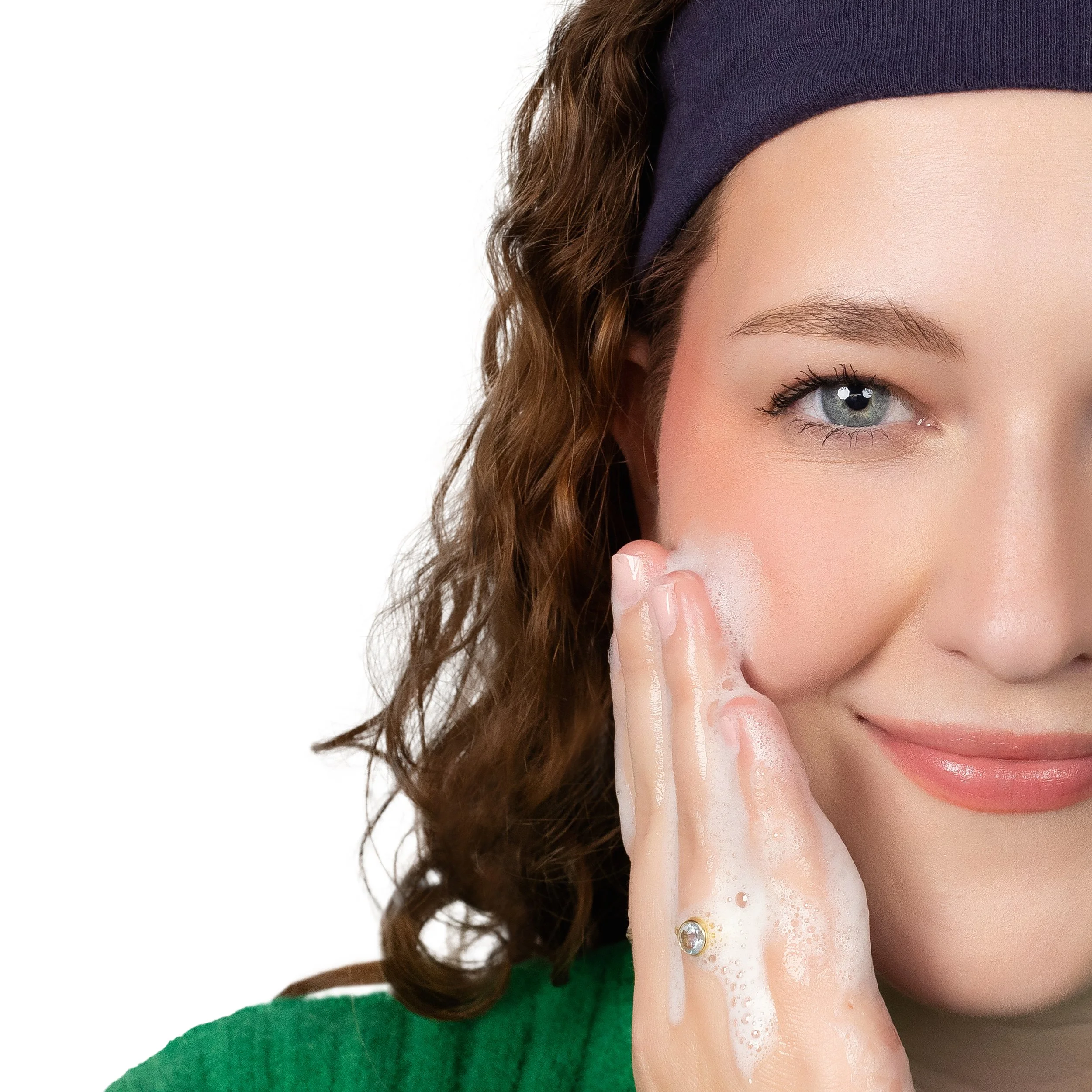 Close-up of a young woman with curly brown hair wearing a blue headband and green shirt, washing her face with soap and water.