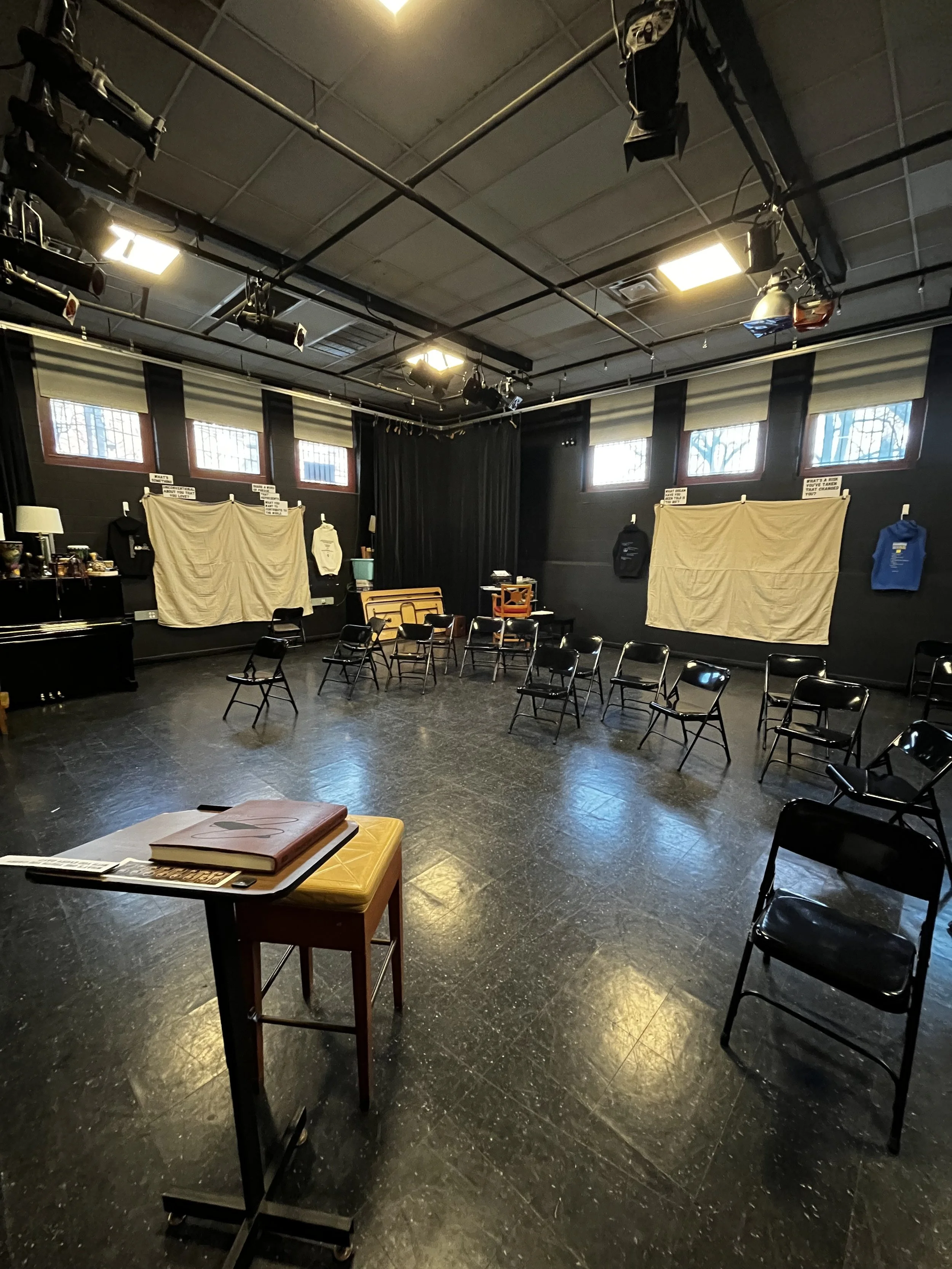 Photo of Black Box theater with textual tapestries hanging from wall, chairs in center, and Living Journal on desk in front