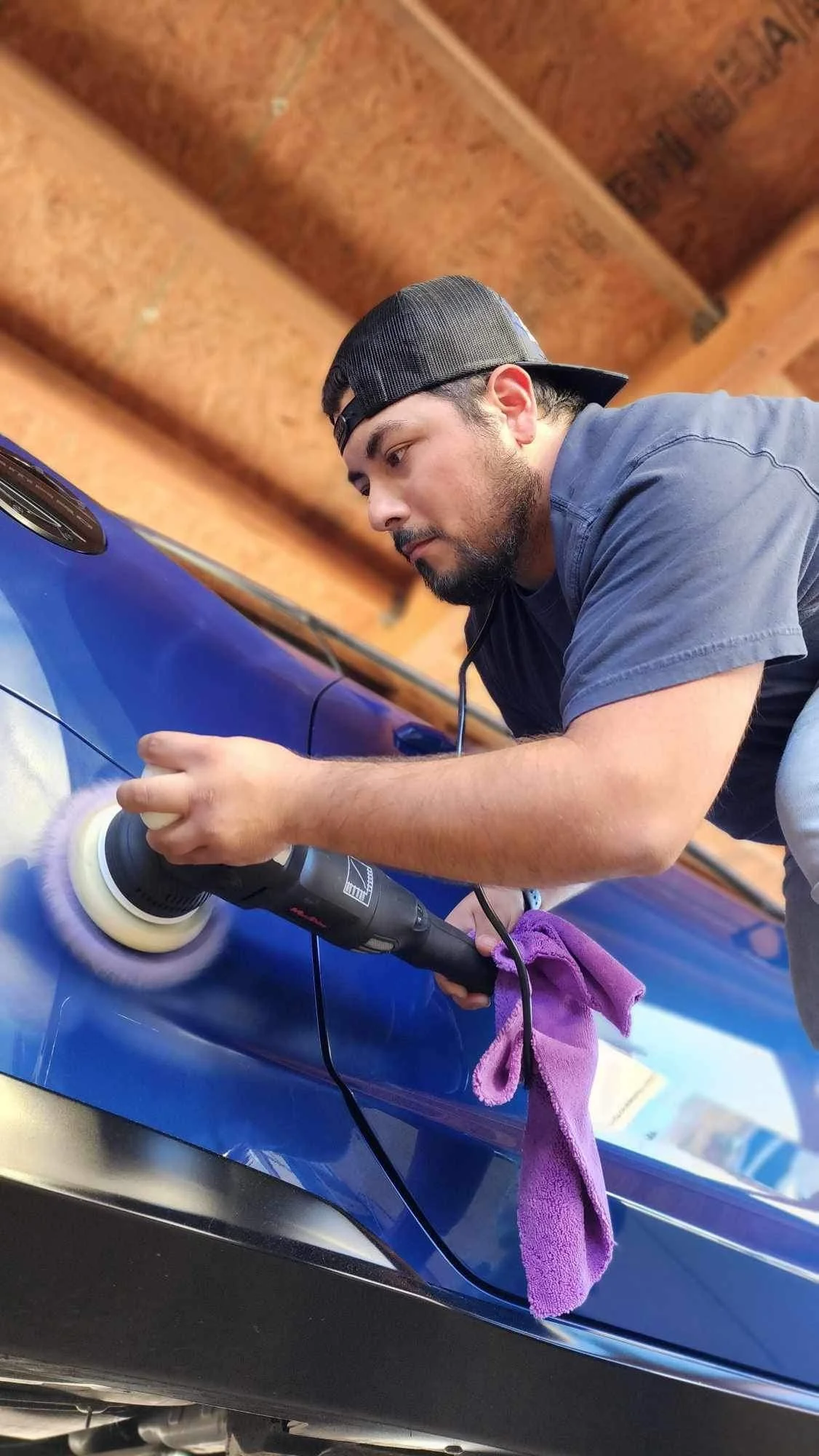 Man polishing a blue car with a buffer, using a purple cloth hanging from his hand.