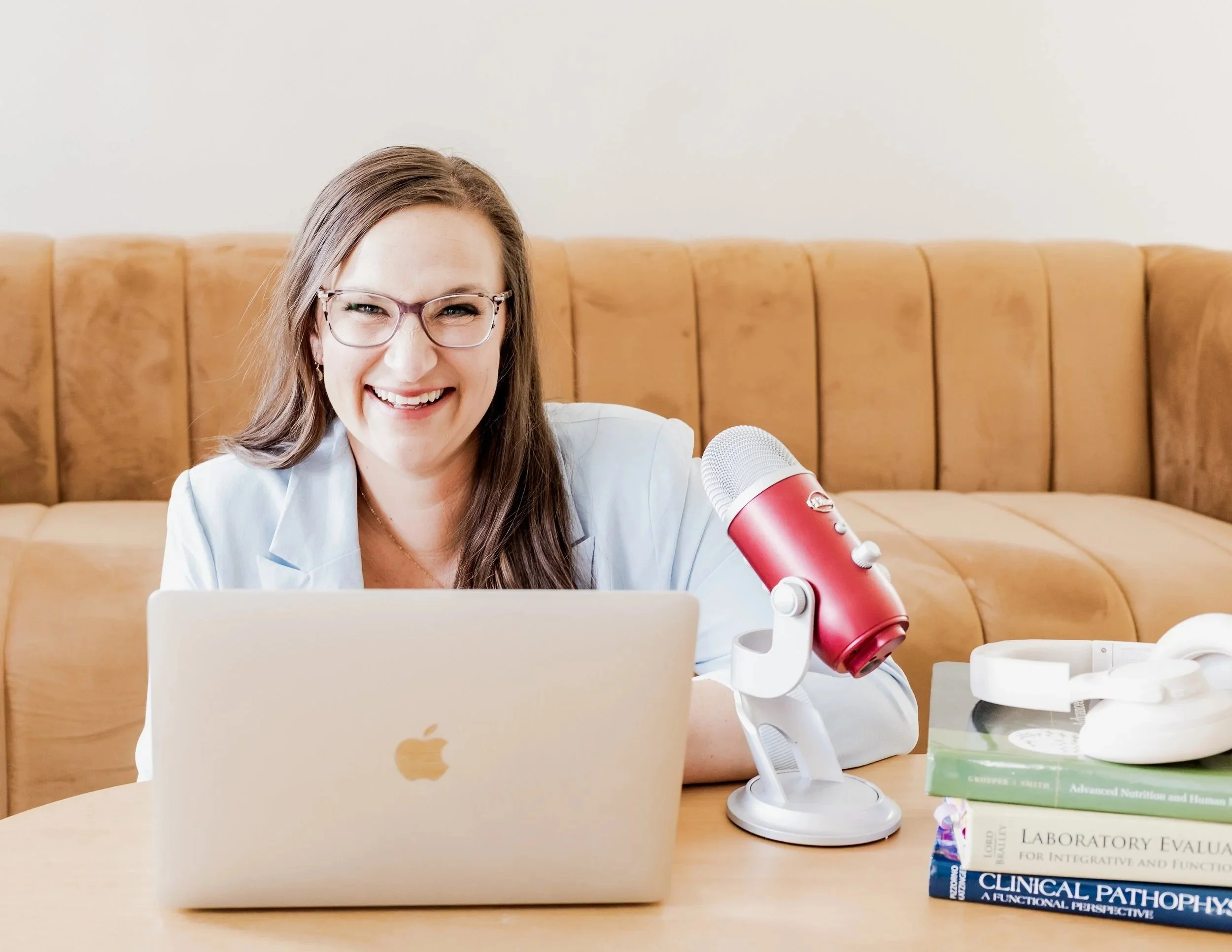 Smiling woman with glasses sitting at a desk with a laptop, microphone, headphones, and health-related books.