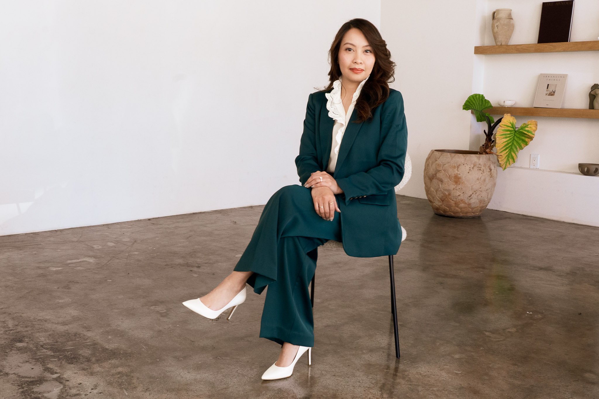 A woman in a teal business suit with white heels sits on a black chair in a minimalist room with a potted plant and wooden shelves on the wall.