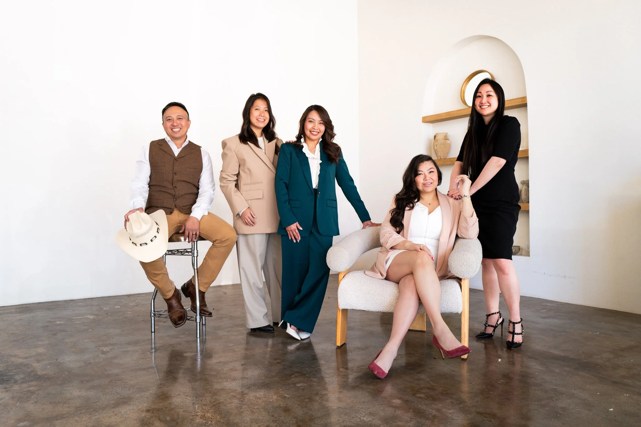 Group of six diverse people dressed in business casual and formal wear posing in a minimalistic indoor space with white walls and wooden flooring.