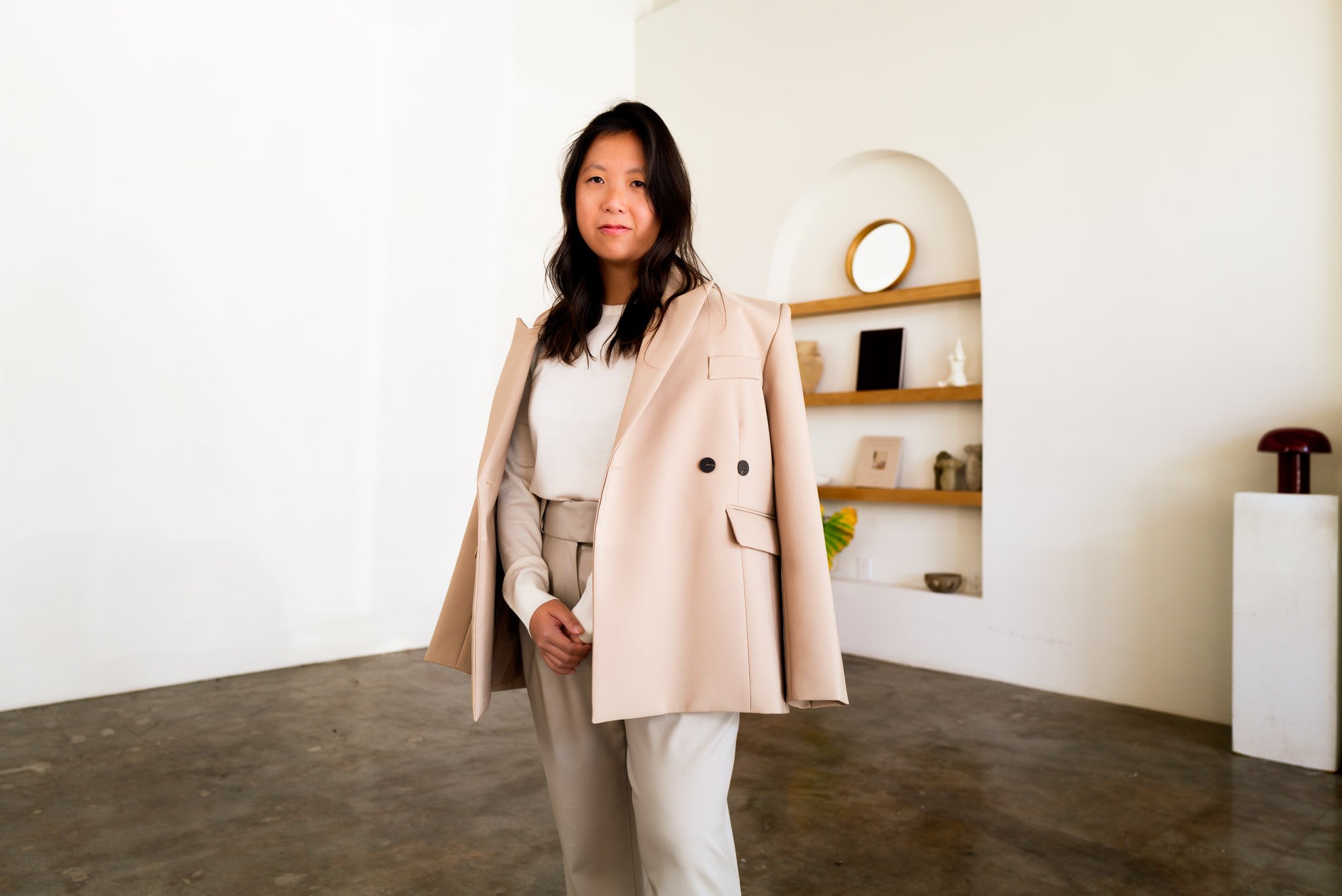 A woman with shoulder-length dark hair stands in a bright, minimalist room with white walls and wooden floor. She is wearing a beige blazer draped over her shoulders, a white blouse, and beige trousers.