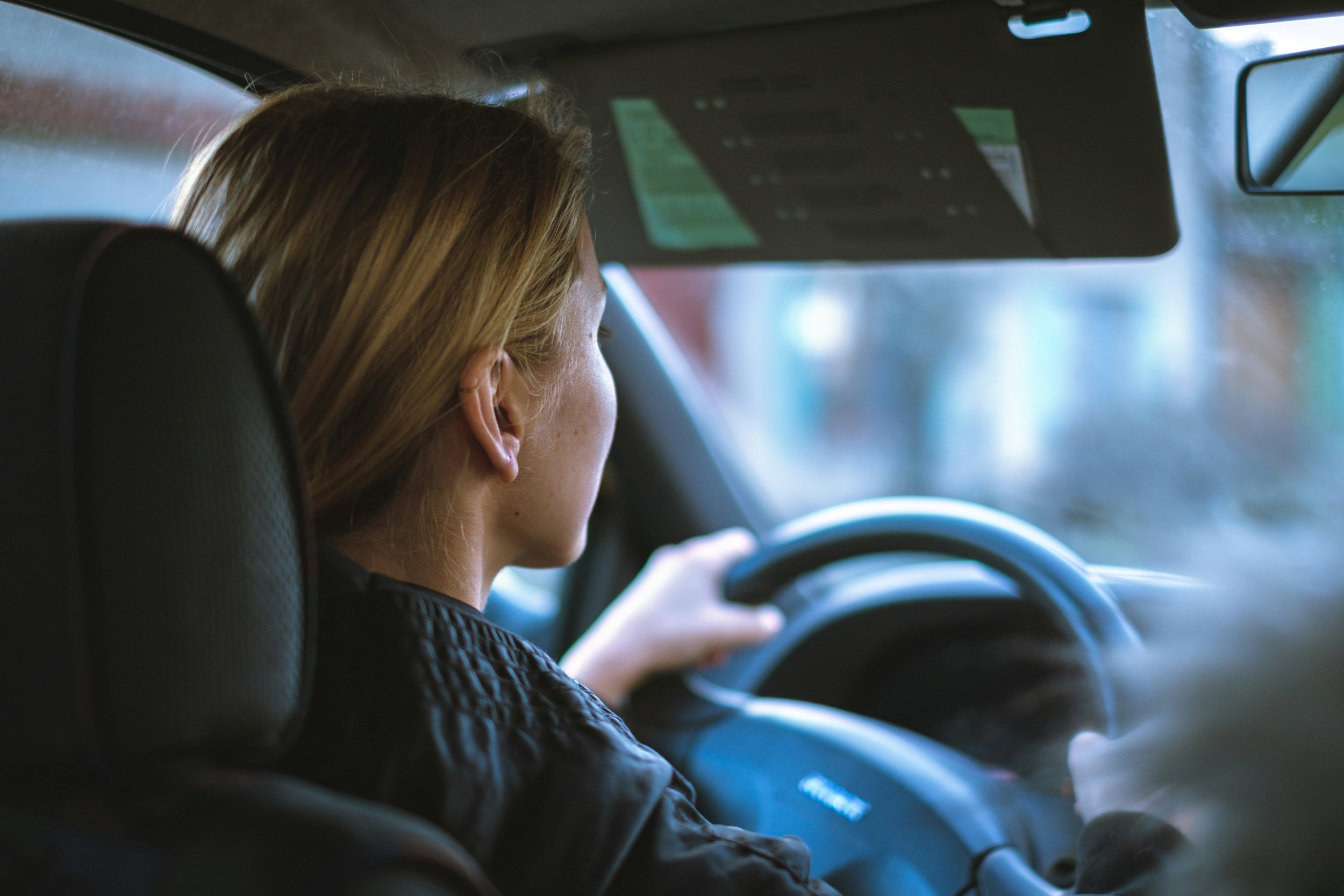 A woman with blonde hair sitting in the driver's seat of a car, holding the steering wheel, seen from the side through the windshield.