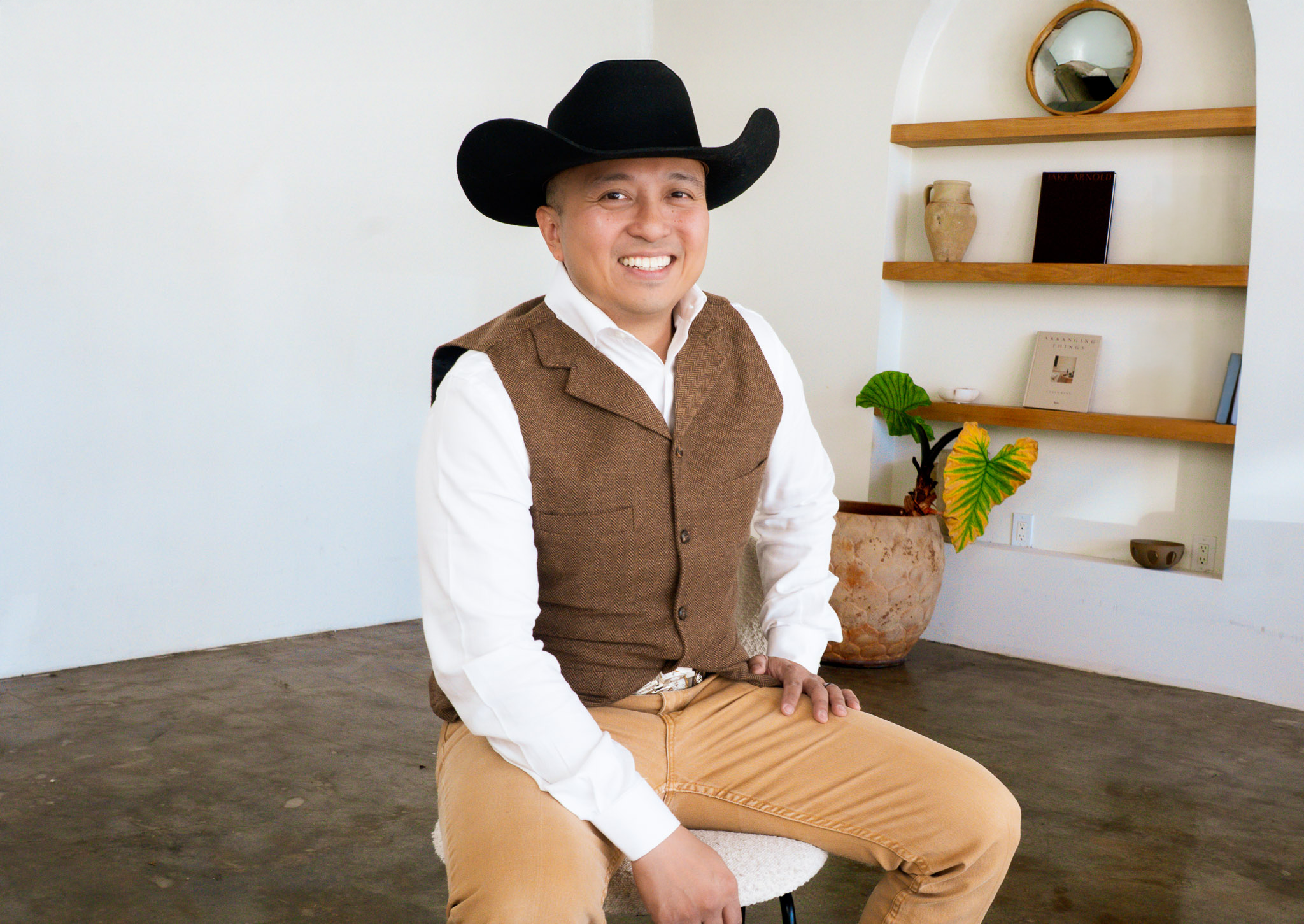 Smiling man in cowboy hat, white shirt, brown vest, and tan pants sitting on a stool in a room with a white wall, wooden floor, and shelves with decorative items and a potted plant.