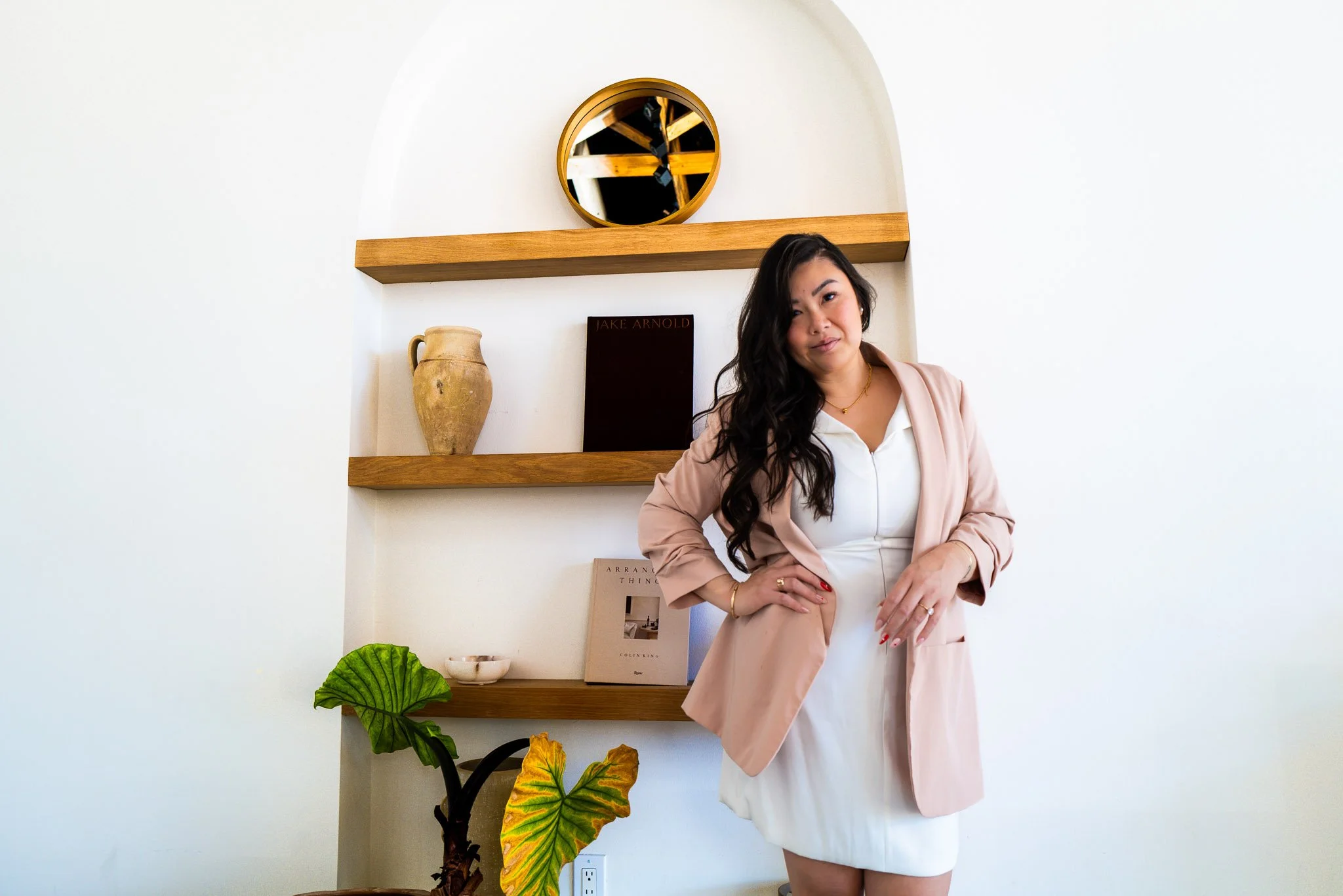 A woman in a white dress and pink blazer standing with her hand on her hip in front of white wall with wooden shelves, decorative vase, books, and a plant.