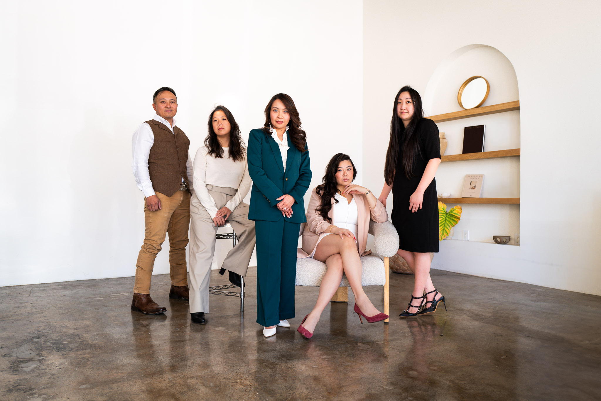 Five diverse individuals in professional attire pose in a minimalistic, modern room with white walls and wooden shelves.