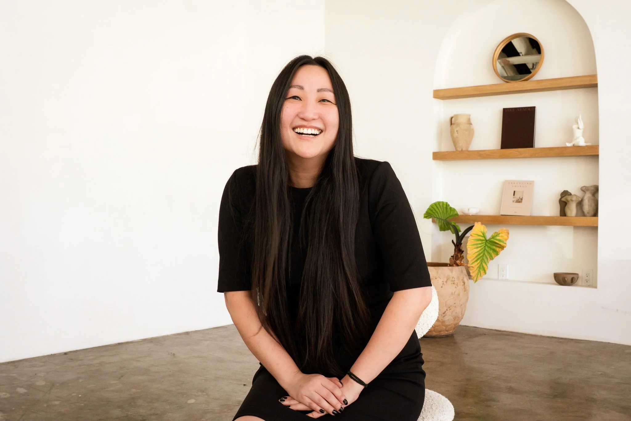 A woman with long dark hair, wearing a black dress, sitting on a white chair and smiling. Behind her is a white wall with wooden shelves holding decorative objects, including a plant, books, and pottery.