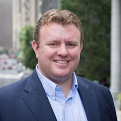 A man smiling outdoors in a blue suit jacket and light blue shirt, with trees and a building in the background.