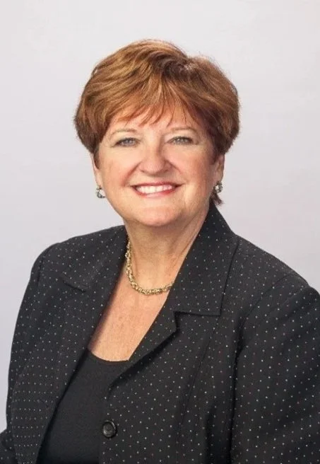 A smiling middle-aged woman with short brown hair, wearing a black blazer with small white dots, gold jewelry, and a black top, against a plain light background.