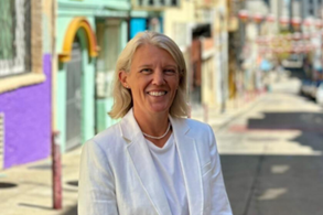A smiling woman with white hair standing on a colorful city street.