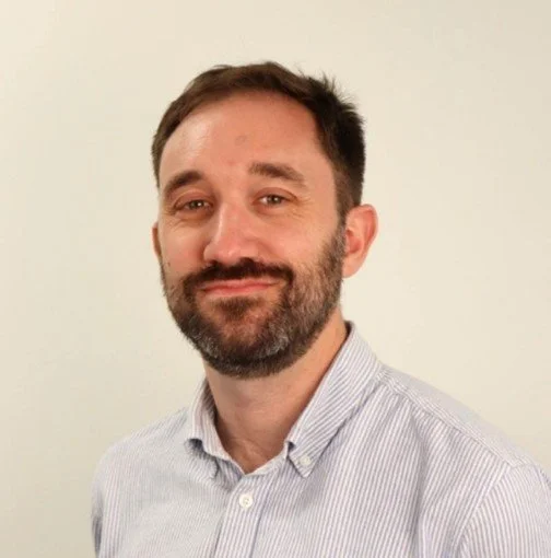 A man with short dark hair and a beard, wearing a light-colored, collared shirt, standing against a plain, off-white background.