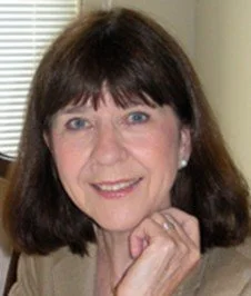Portrait of a woman with shoulder-length brown hair, smiling, wearing a beige top, with a hand resting near her chin.