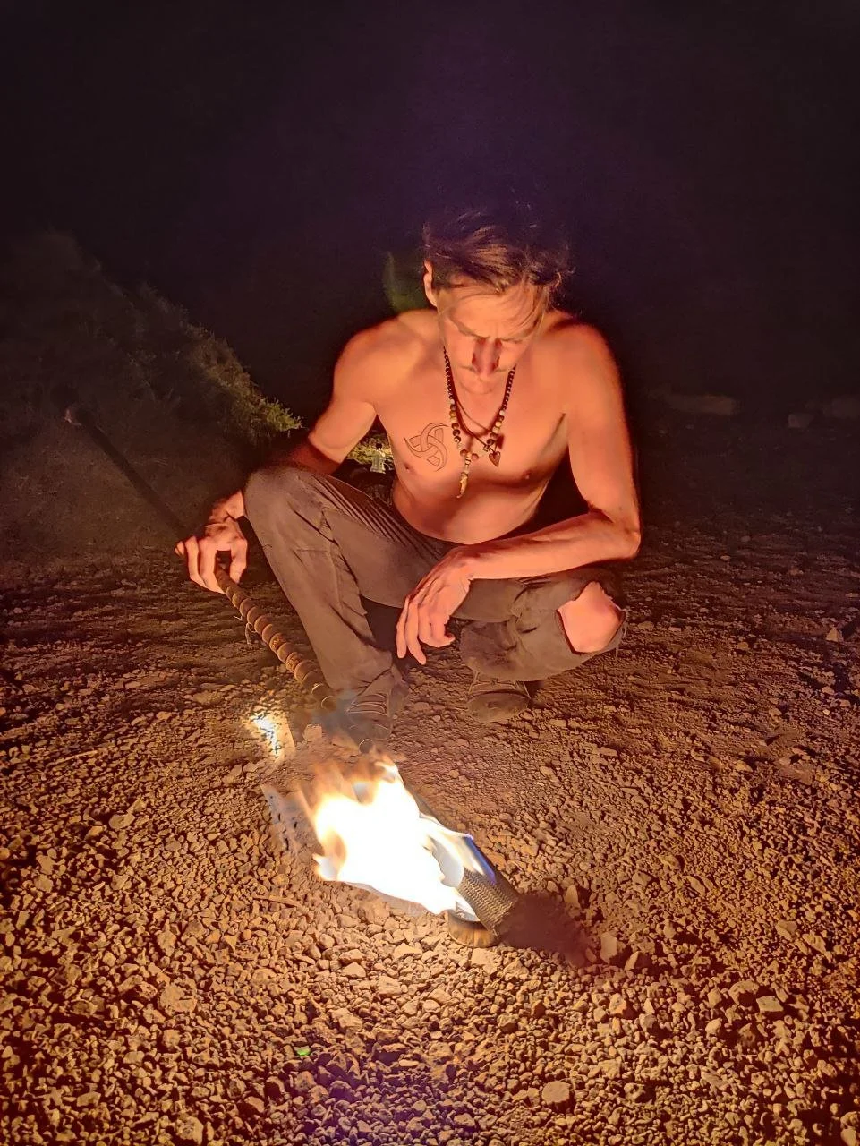 Ein nackter Mann mit langen Haaren und mehreren Halsketten sitzt nachts auf einer erdigen Oberfläche und hält eine brennende Feuerkeule.