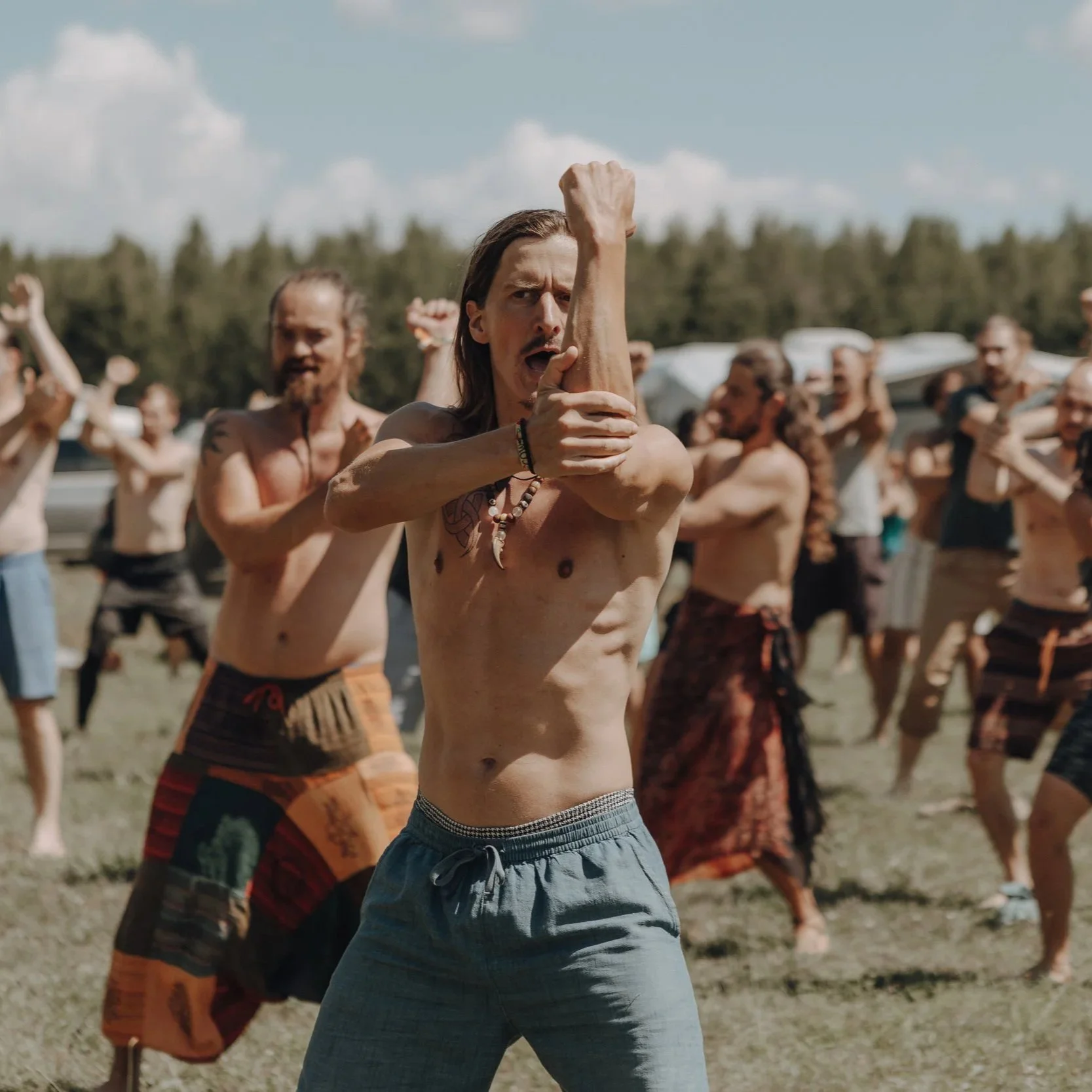 Mehrere hervortretende Männer, die an einem Outdoor-Festival teilnehmen, tanzen Haka und bewegen sich frei, mit einer Naturkulisse im Hintergrund. Simon Harm Brunken Schamane AUFSTELLUNGEN, HAKA KRAFTTANZ & SCHAMANISCHE RITUALE