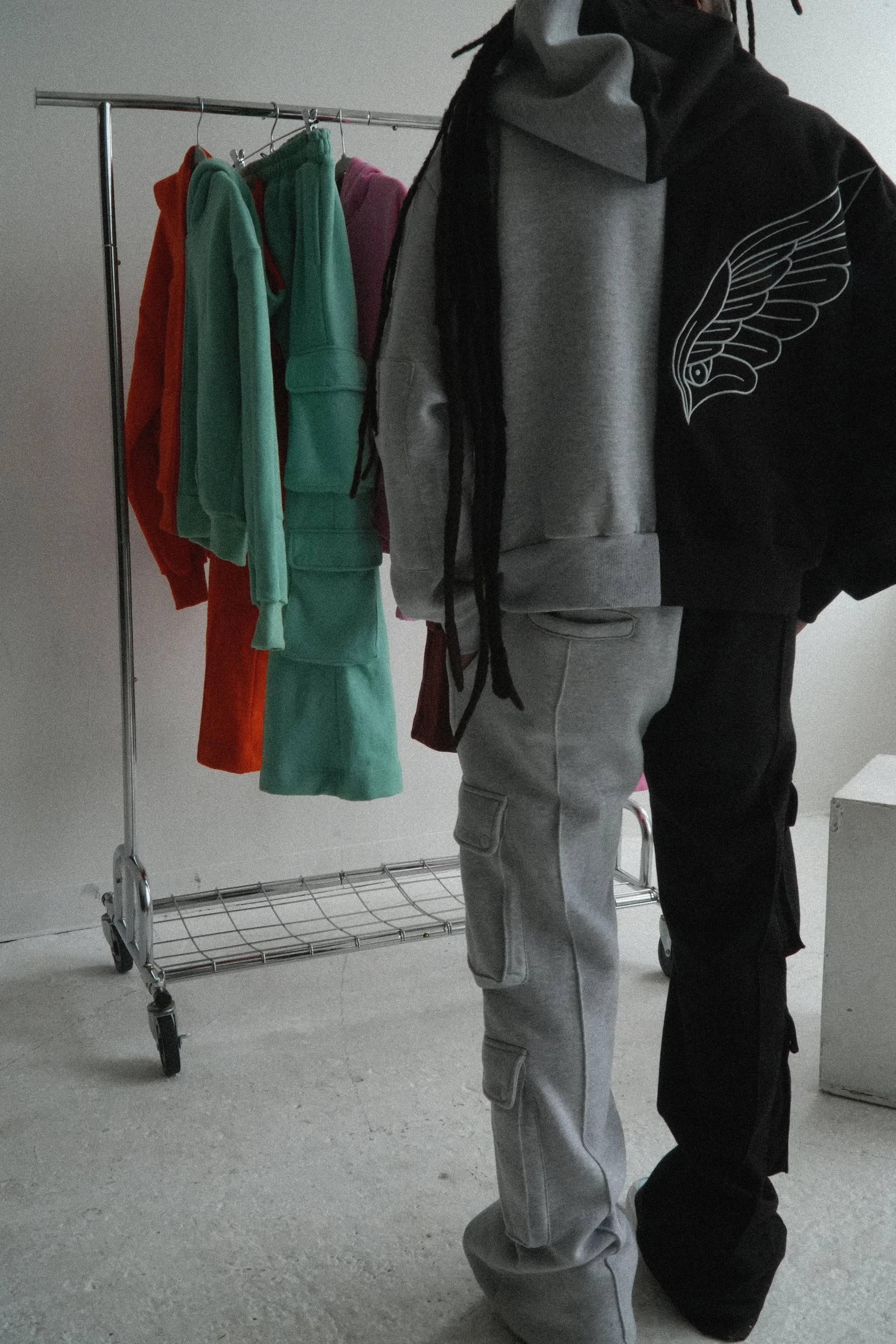 rapper and surrealist artist chester watson / monti in front of a clothing rack with a collection of heavyweight hoodies hanging on it. the hoodie and sweatpants are both half grey and half black cut and sew construction