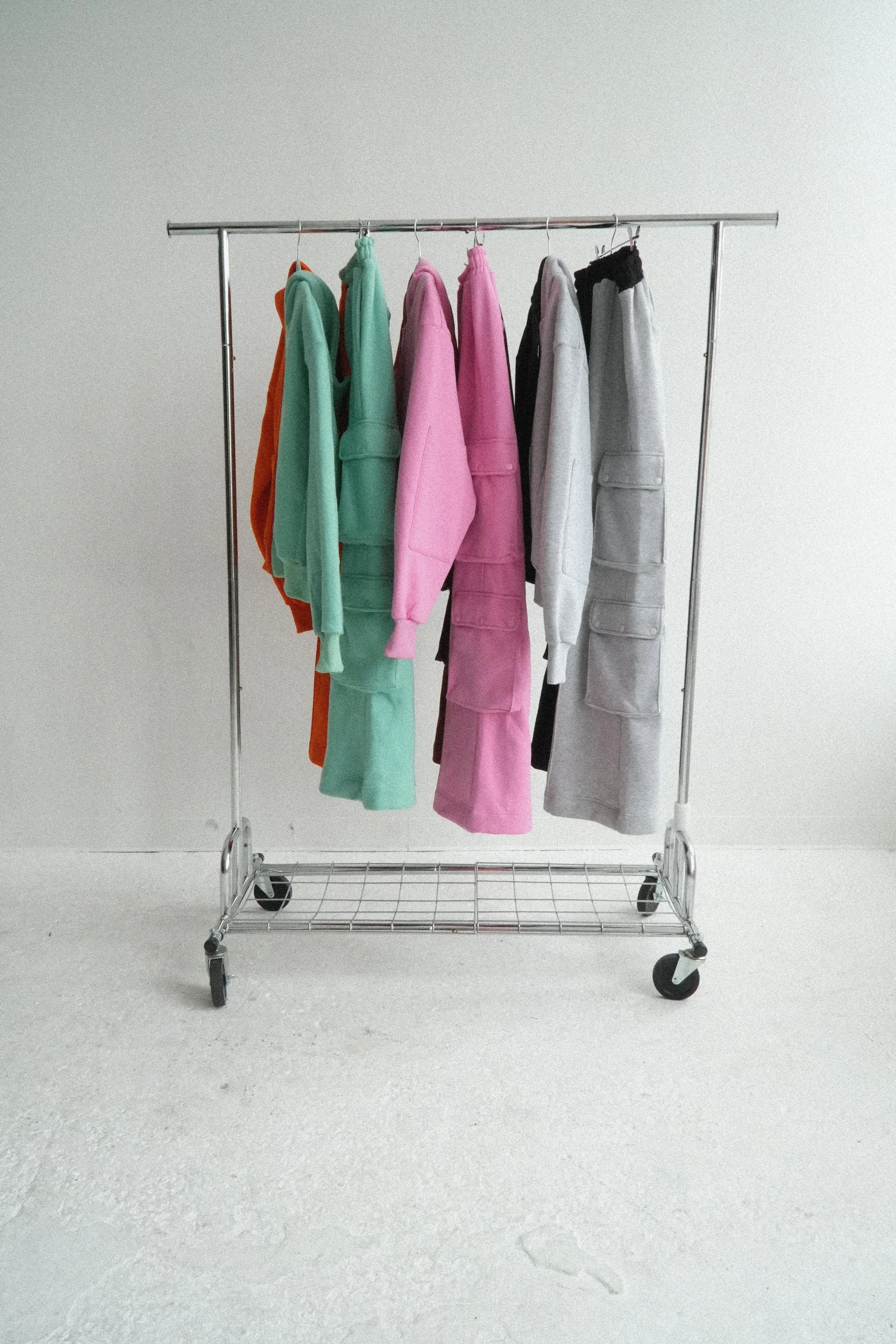 a collection of pastel and neutral-colored sweatshirts and sweatpants including orange, seafoam, pink, brown, black, and grey against a plain white wall hanging from a metal clothing rack with wheels