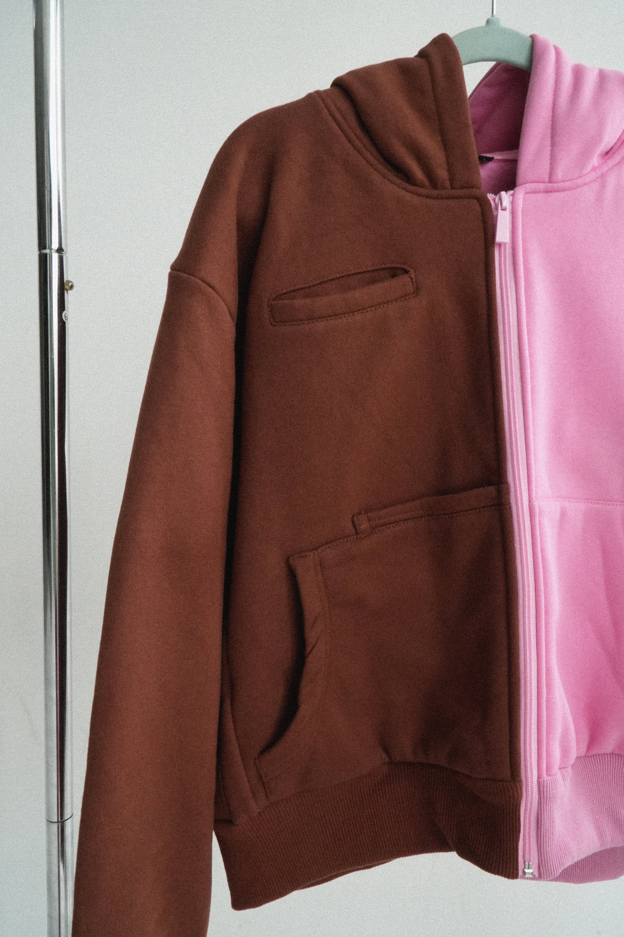 A unique half and half brown and pink zip-up hoodie hanging on a hanger. the clothing is of high heavyweight quality reflecting luxury goods
