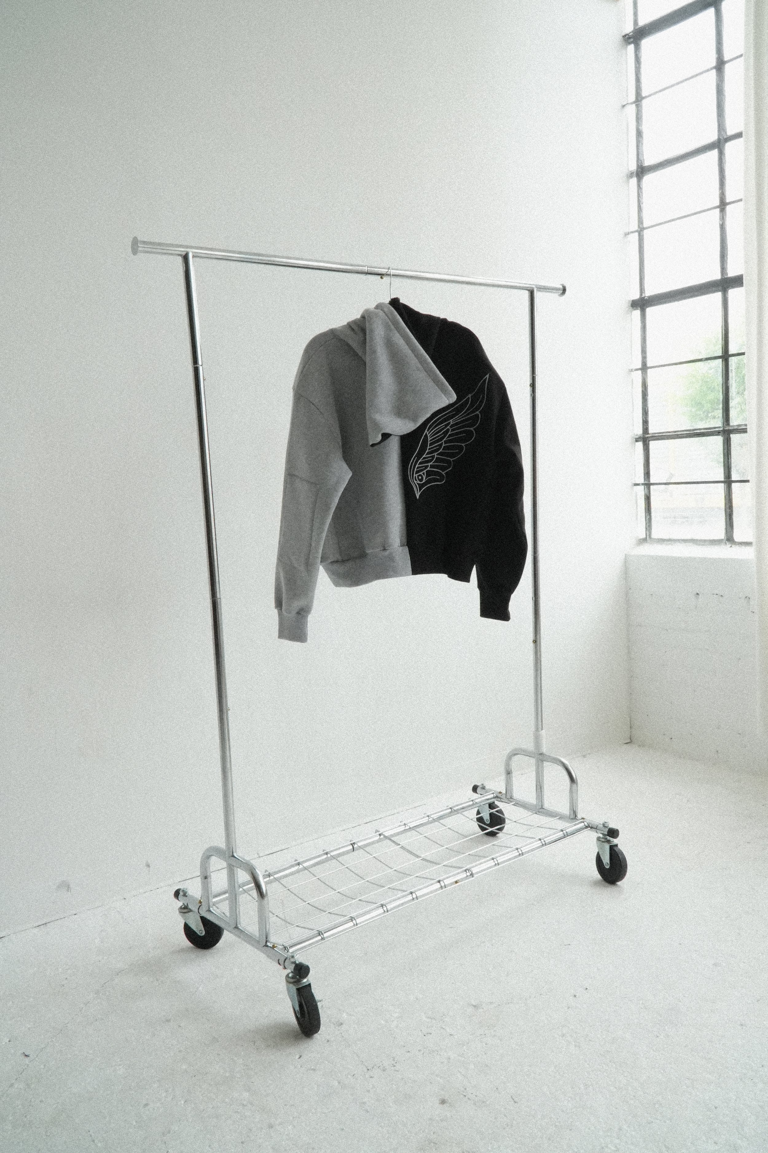 A clothing rack on wheels holding one half grey / half black hooded sweatshirt with a large white wing design embroidered on the backside, set against a white wall and a window with multiple panes in a minimalist industrial room