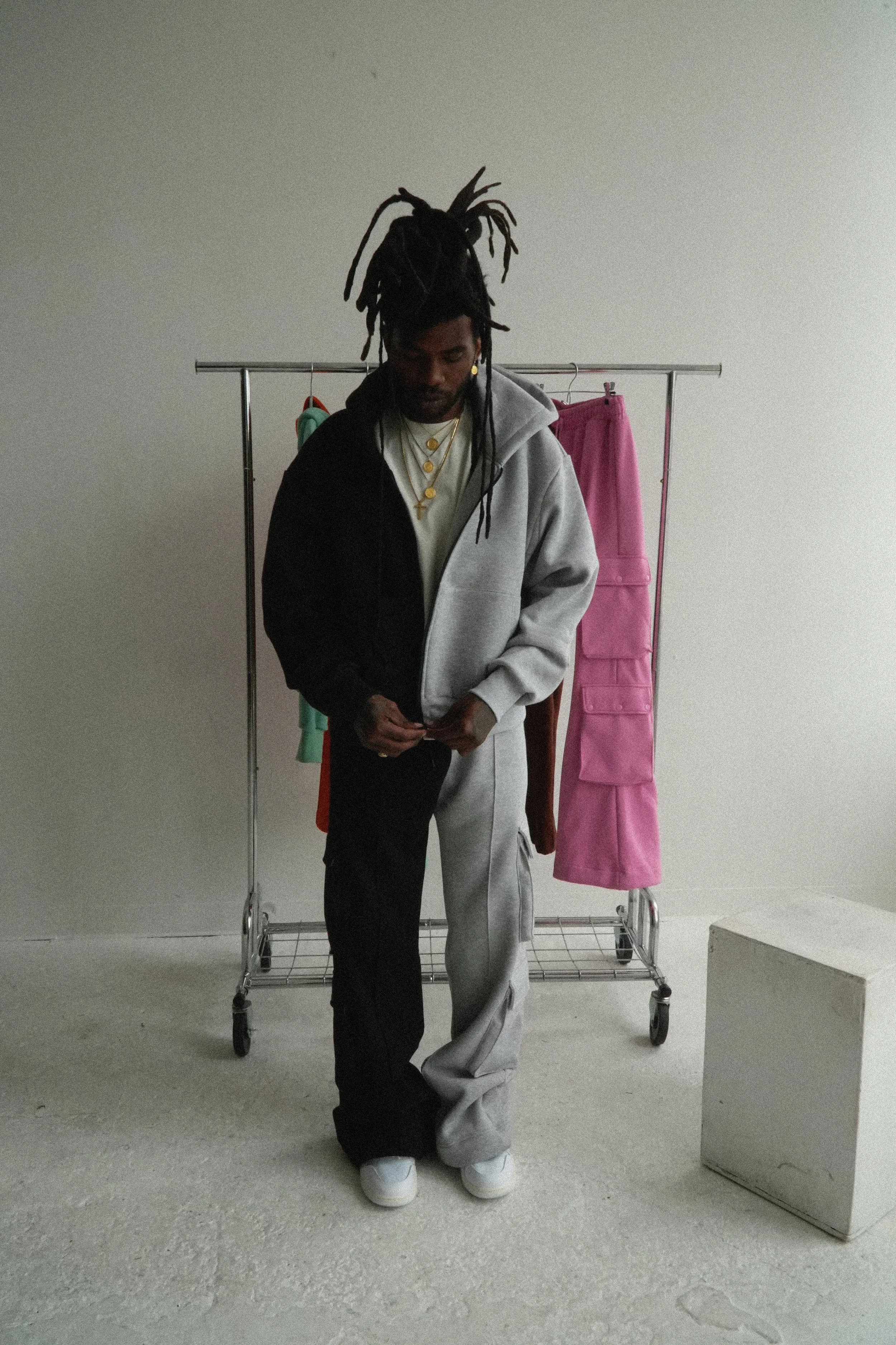 rapper and surreal artist chester watson / monti with dreadlocks standing in front of a clothing rack with colorful garments, wearing a gray and black hoodie, white sneakers, and layered gold necklaces.