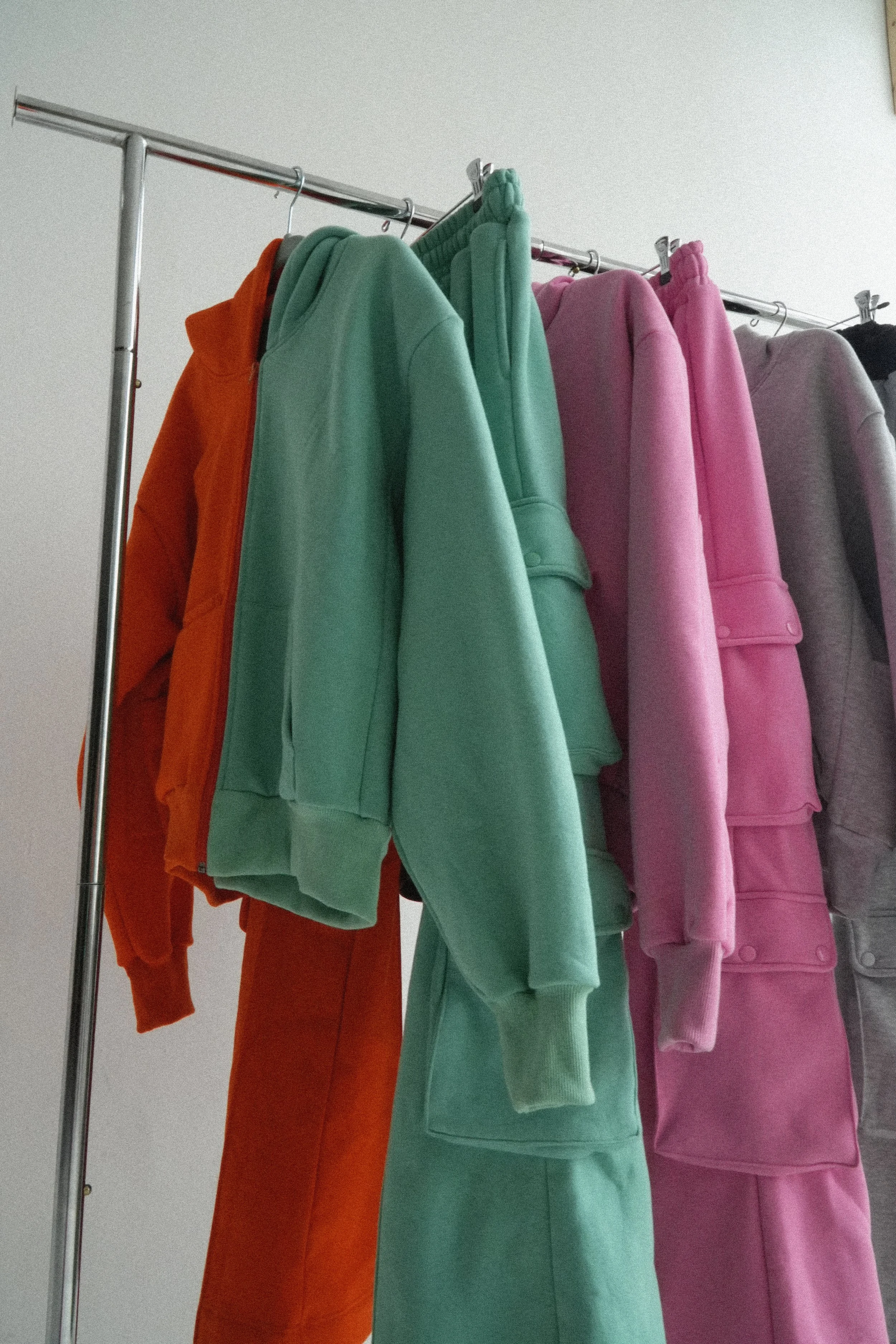 a collection of colorful sweatshirts and sweatpants hanging on a metal clothing rack, including orange, seafoam, brown, pink, grey, and black. the clothing is of high heavyweight quality reflecting luxury goods