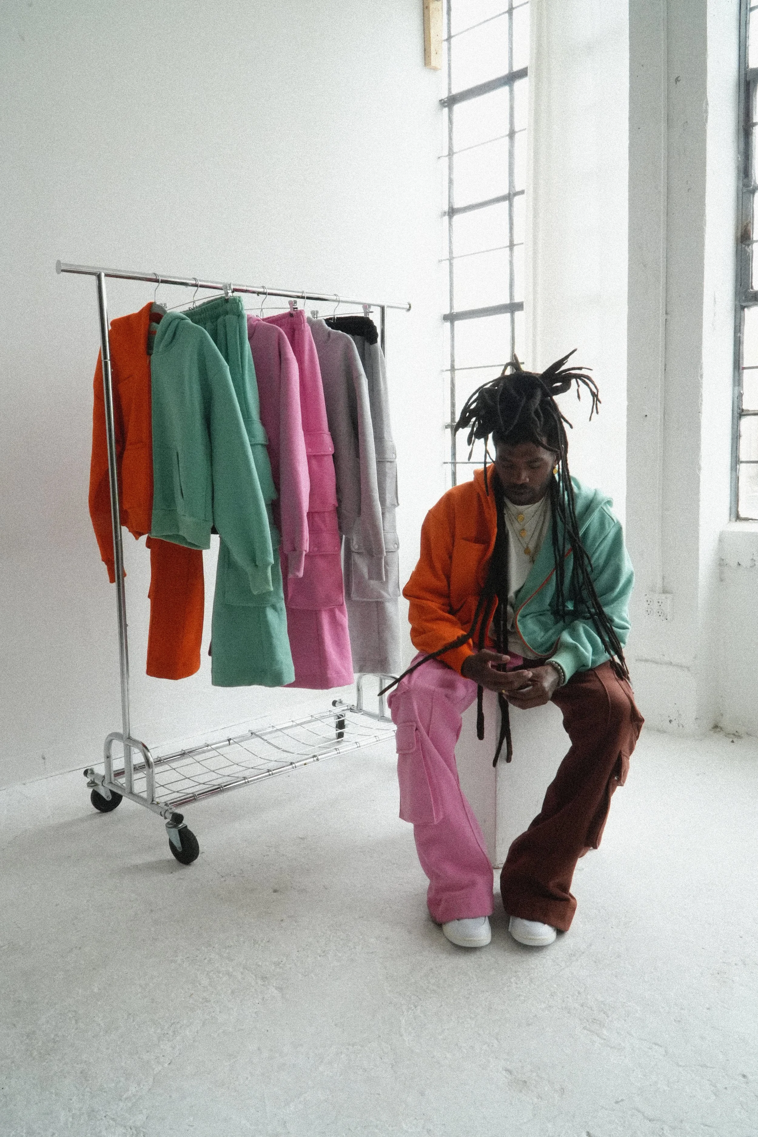 rapper and surrealist artist chester watson / monti with long dreadlocks is sitting on a stool in a minimal industrial-style room with white walls and large windows. next to him is a clothing rack with a collection of pastel-colored hoodies and pants