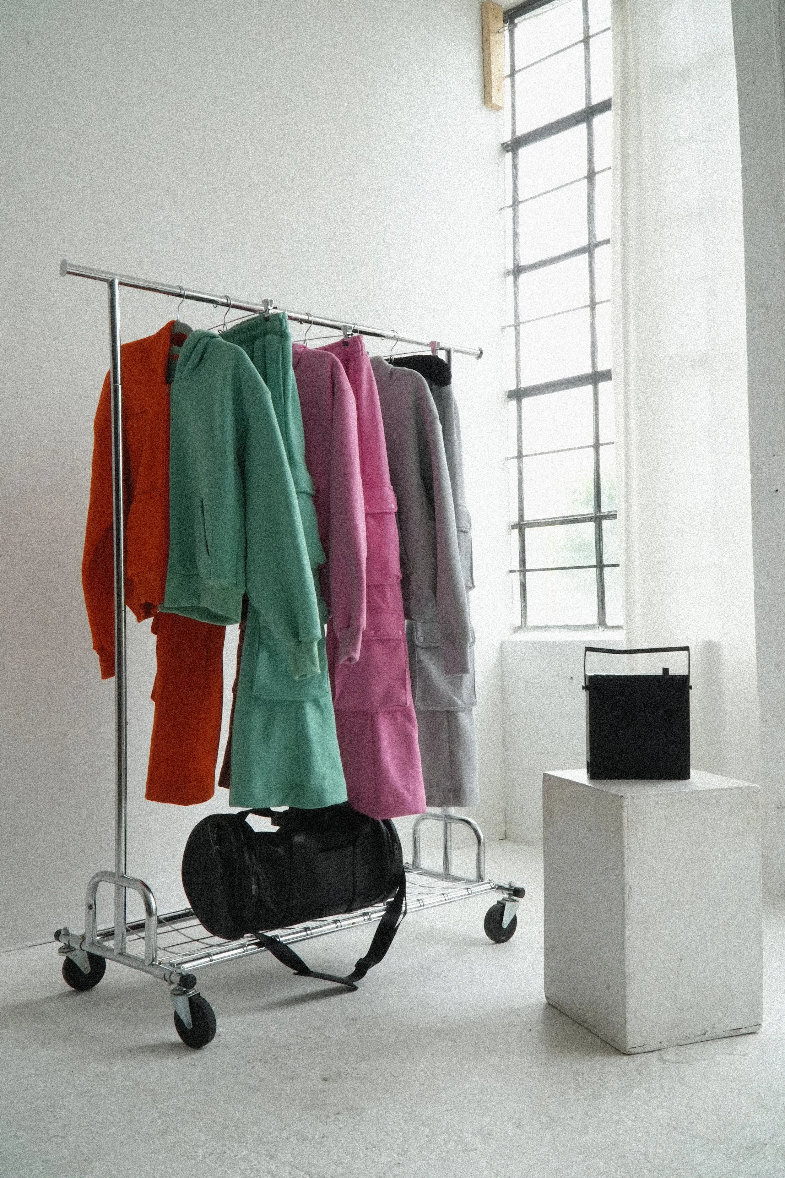 high quality pastel colored sweatsuits hanging on a metal clothing rack inside a minimalist industrial room with a black leather i love ugly bag under the rack and a black teenage engineering ob-4 speaker on a white pedestal nearby