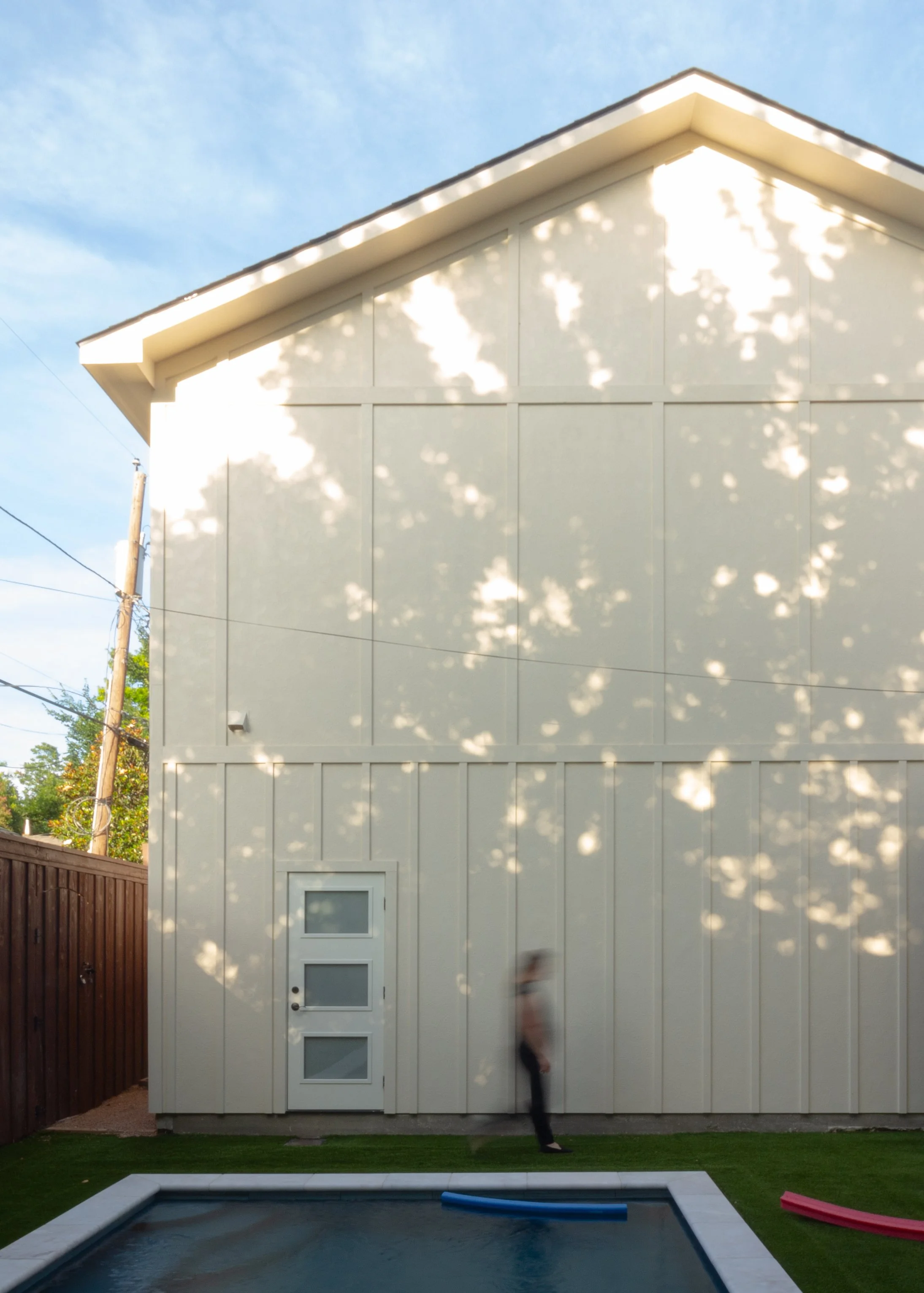 Two-story back house with off-white exterior walls and a modern door with glass panels. Shadows of tree leaves are cast across the building facade. There is a small pool in the foreground and a person walking near the house. The sky is blue.