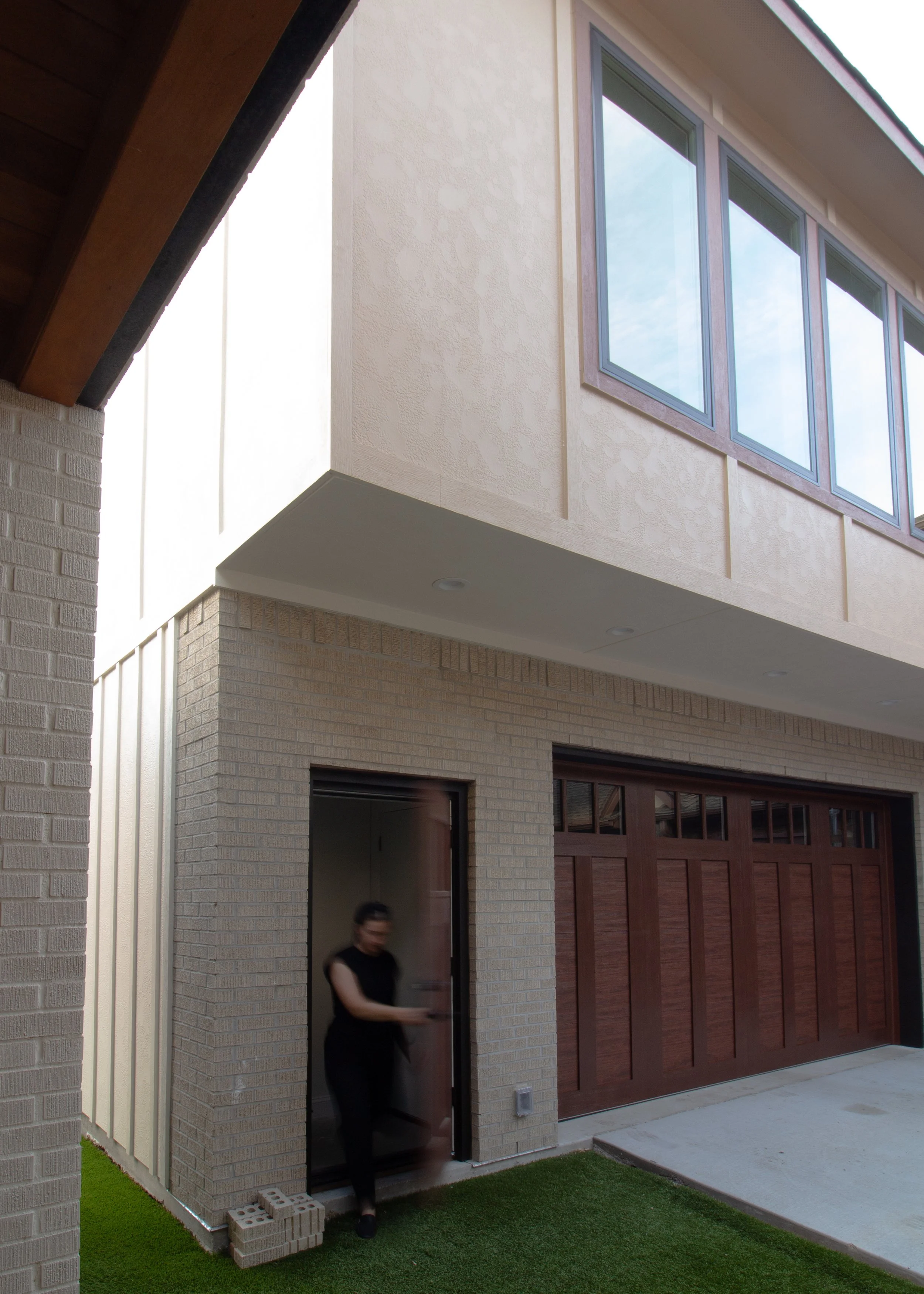 A person walking out of a doorway of a back house with a brick wall, wooden garage door, and large windows on the upper level.