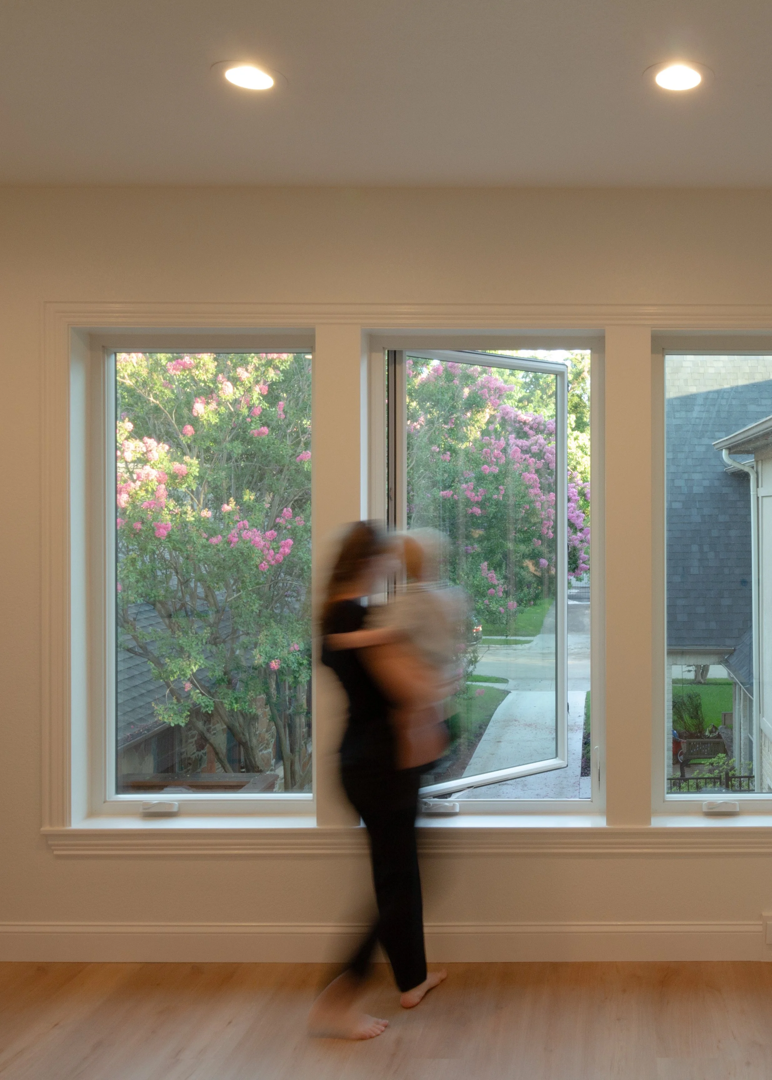 A person holding a child standing by a large window with an open window pane showing pink flowering trees outside, in a room with wooden floors, white walls, and ceiling downlights.