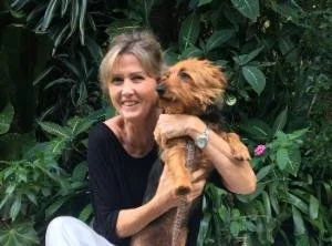 A woman holding a small brown dog outdoors surrounded by green foliage.