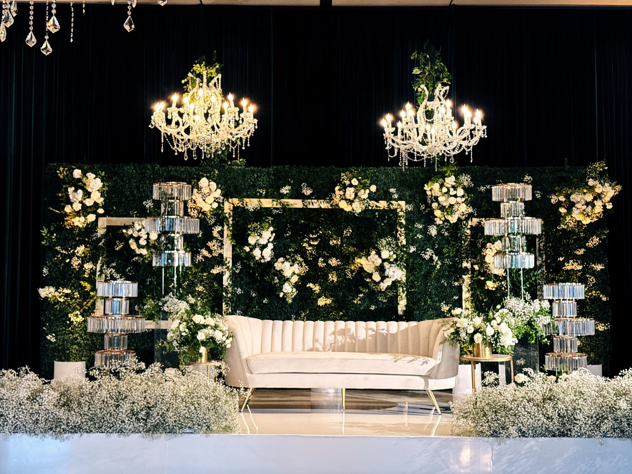 Elegant wedding or event stage decorated with white roses and greenery, two chandeliers hanging from above, white velvet sofa in the center, surrounded by tall crystal candle holders and floral arrangements.